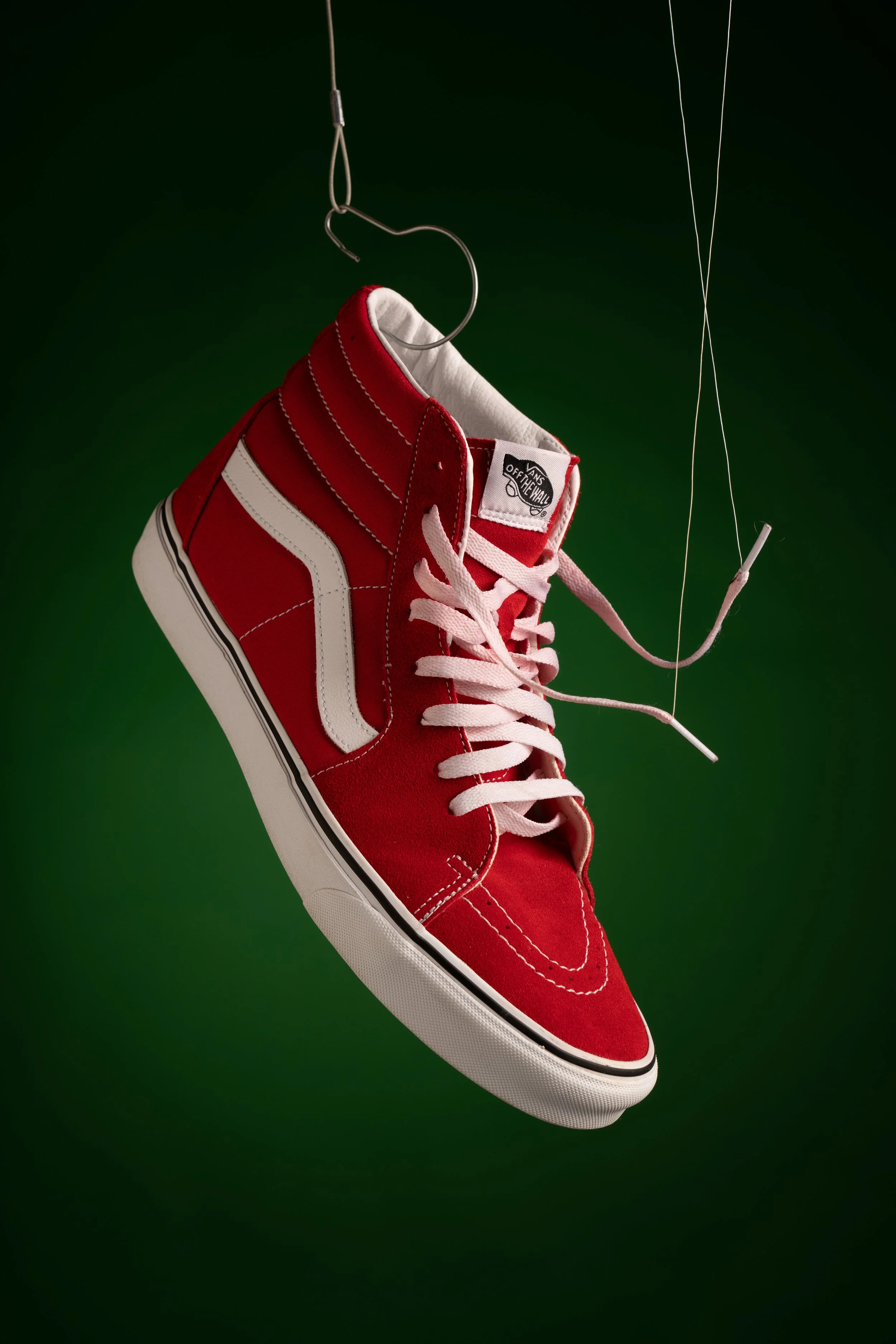 Red and white Vans sneaker hanging from a wire against a dark green background.