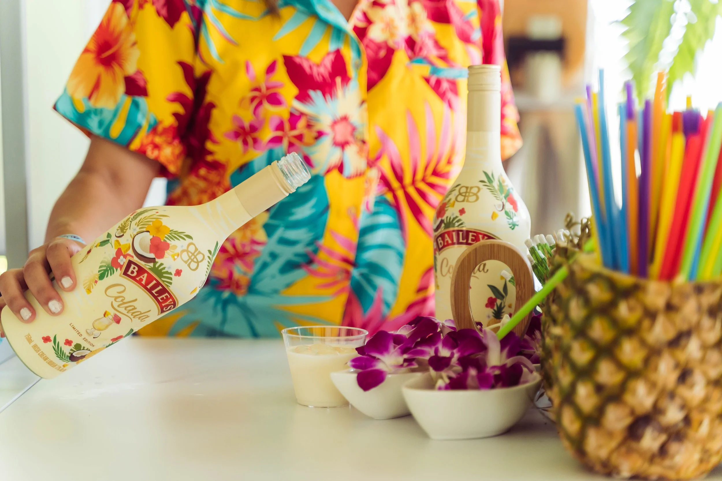 A person in a colorful Hawaiian shirt pouring Bailey's Irish Cream from a bottle into a glass, with tropical decorations, fruit, and straws on a white table.