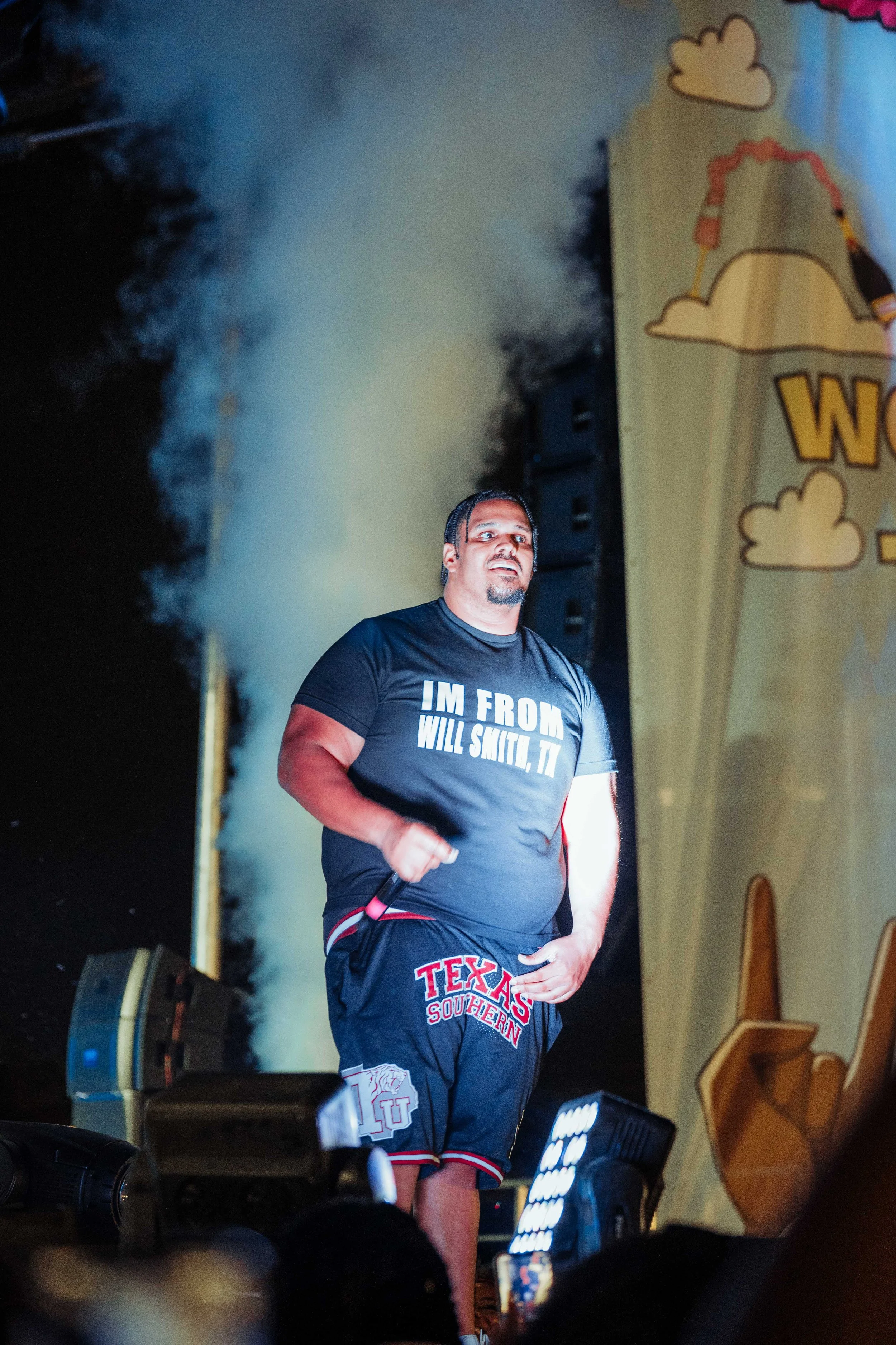 A man standing on stage during an event, wearing a black T-shirt that says 'I'M FROM WILL SMITH, TI' and black shorts with Texas Southern University logo, with smoke or fog behind him and large speaker on stage.