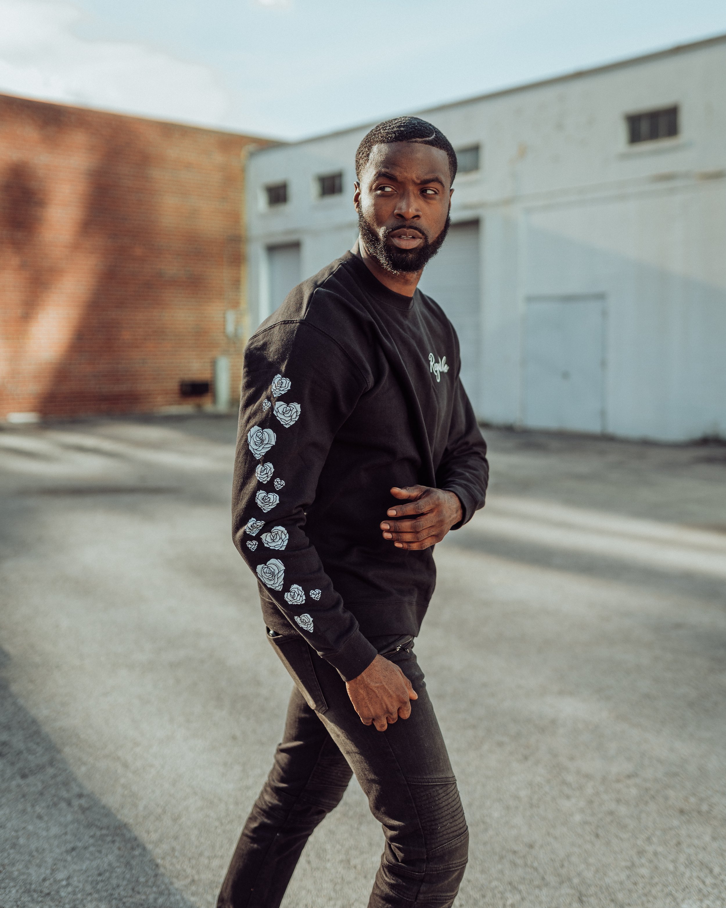 A young man with a beard, wearing a black sweatshirt with floral designs on the sleeves, standing outdoors on a paved area with a white building and brick wall in the background.