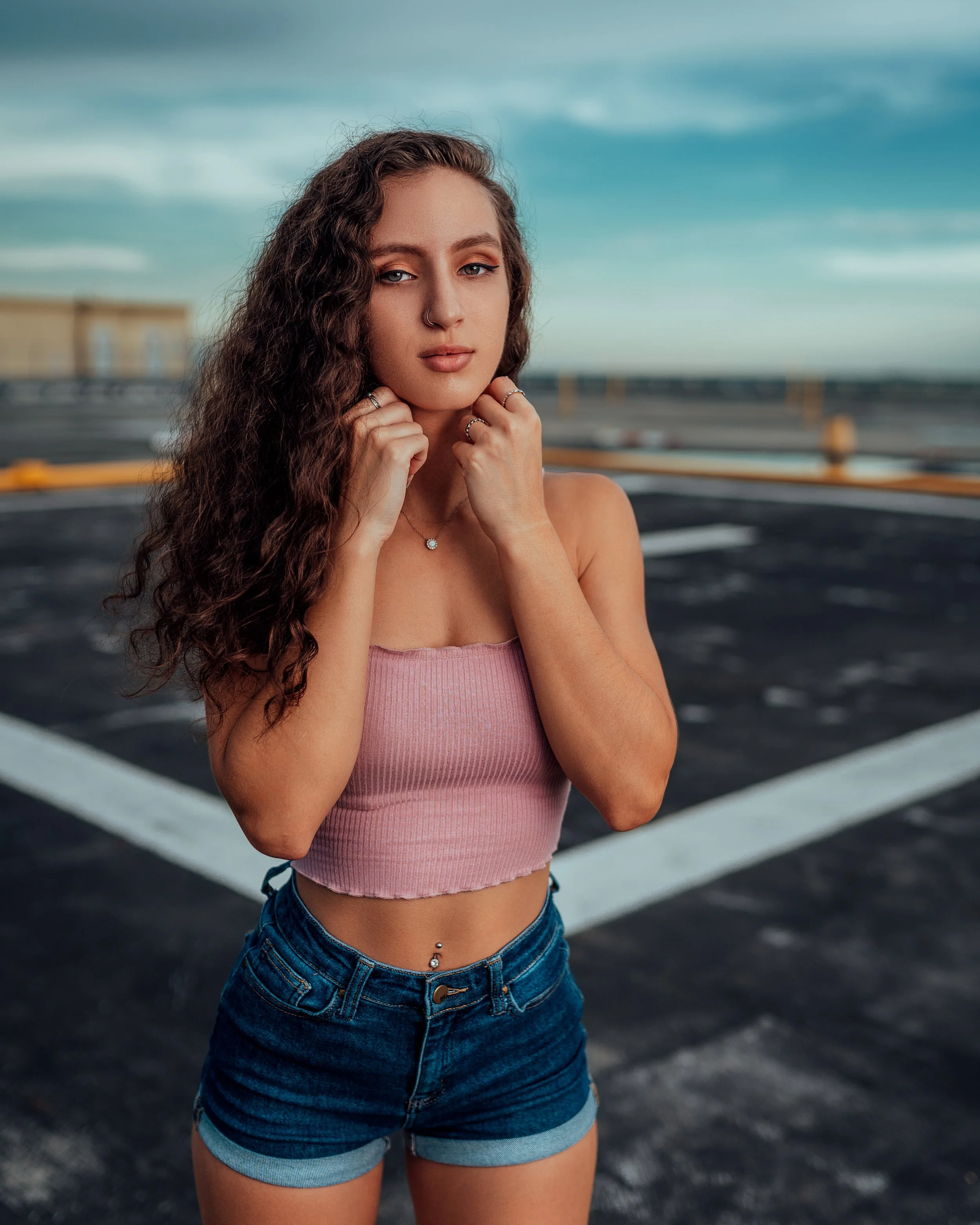 A young woman with long curly hair standing in an empty parking lot, wearing a pink crop top and denim shorts.