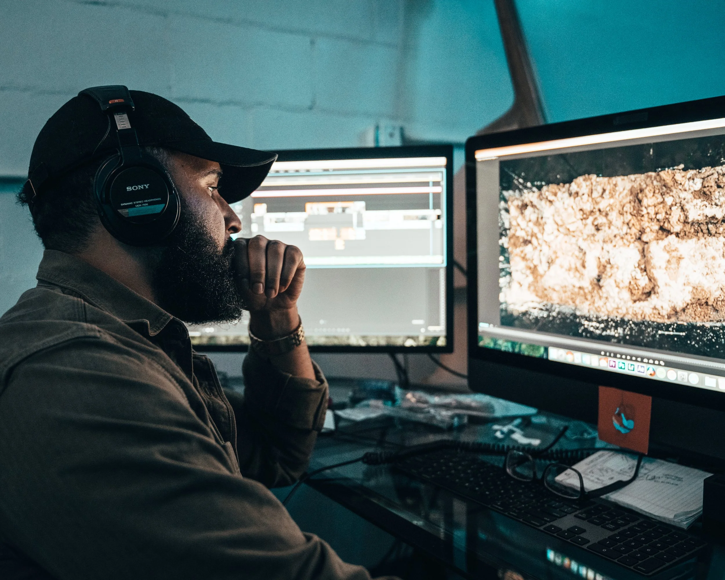 Man with beard wearing headphones and cap working on digital art or video editing at desk with dual monitors, one displaying a soil or earth image.