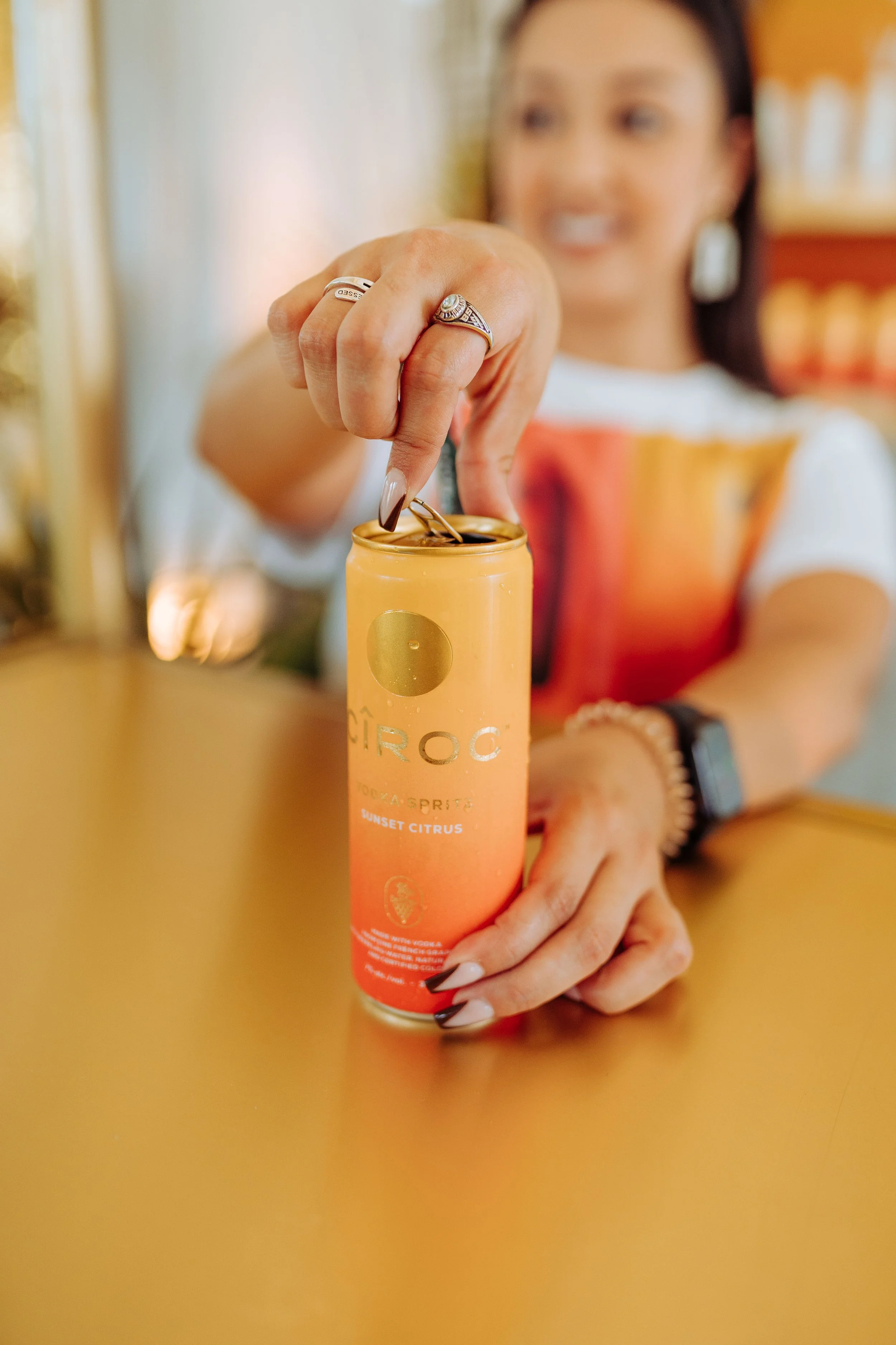 A woman with long dark hair and jewelry opening an orange and yellow can of Ciroc Sunset Citrus vodka.