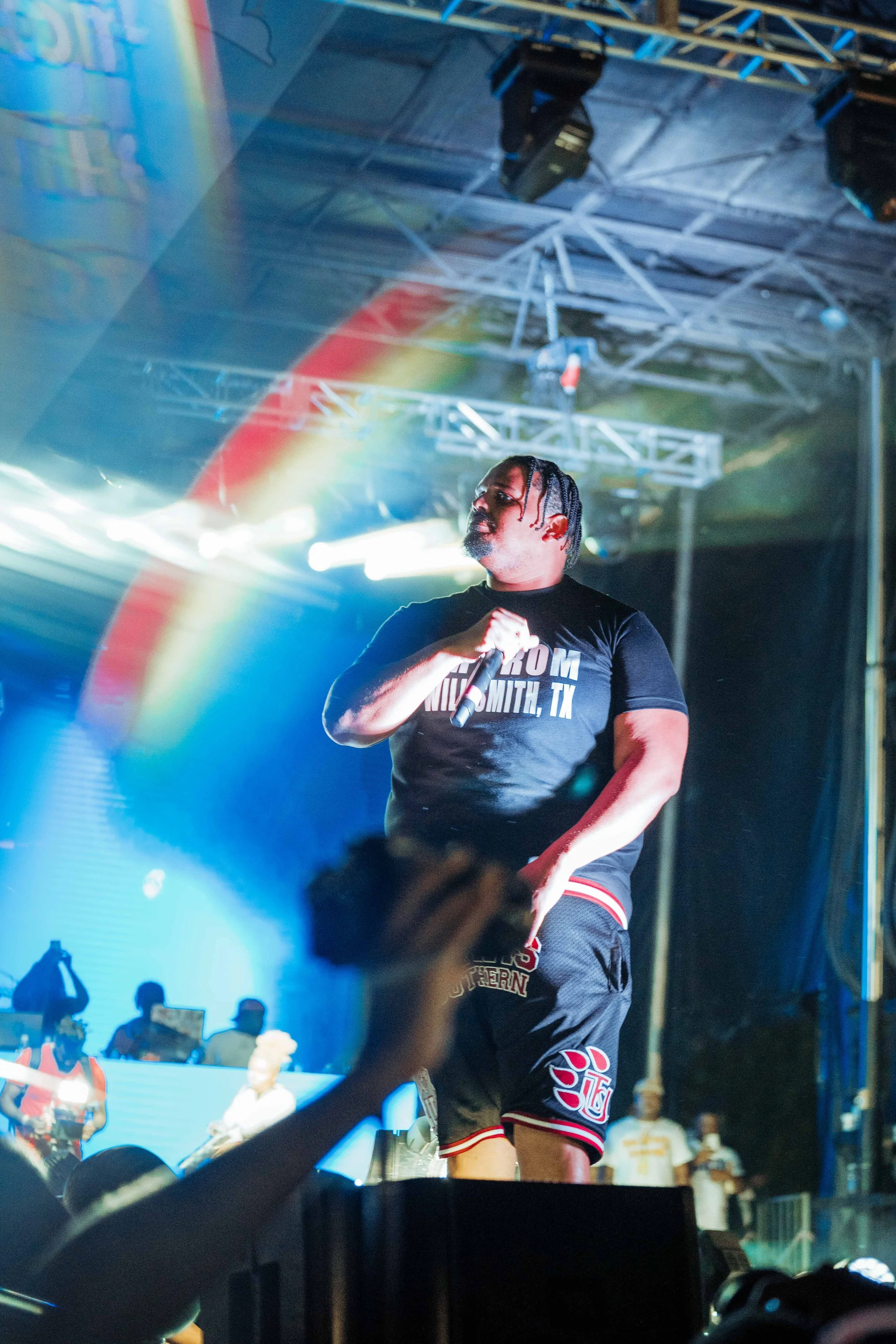 A male performer is on stage holding a microphone with colorful stage lights and people in the audience in the background.