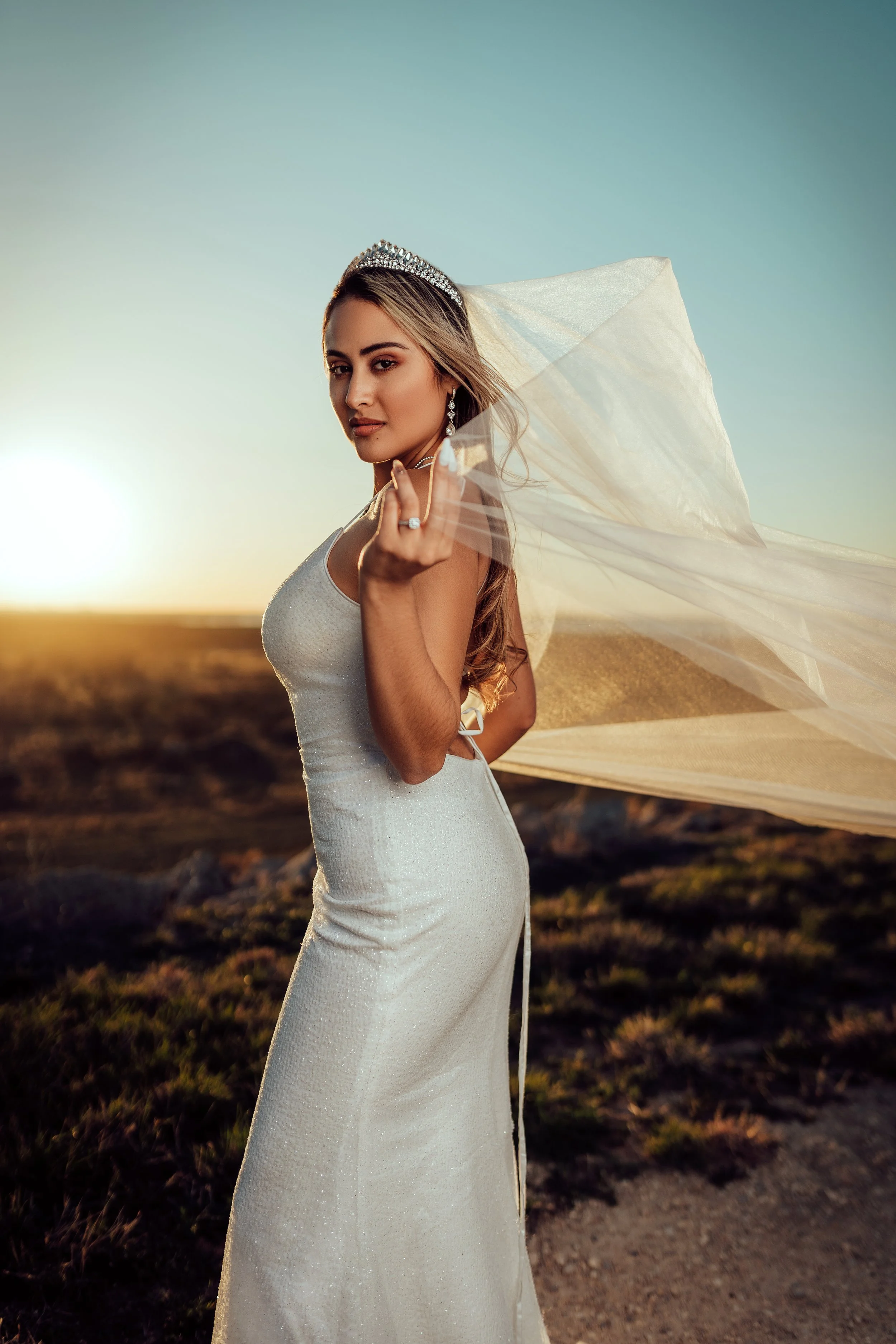 A woman in a white wedding dress with a veil outdoors at sunset, wearing a tiara and jewelry, looking at the camera.