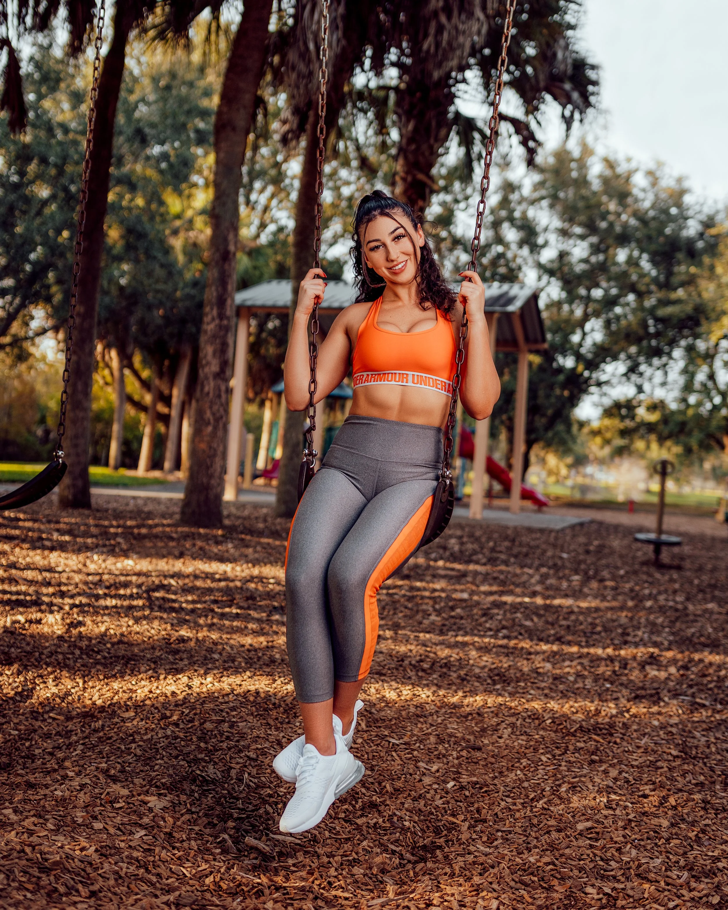 A young woman sitting on a swing at a park during daytime, smiling at the camera, wearing an orange sports bra, gray and orange leggings, and white sneakers.