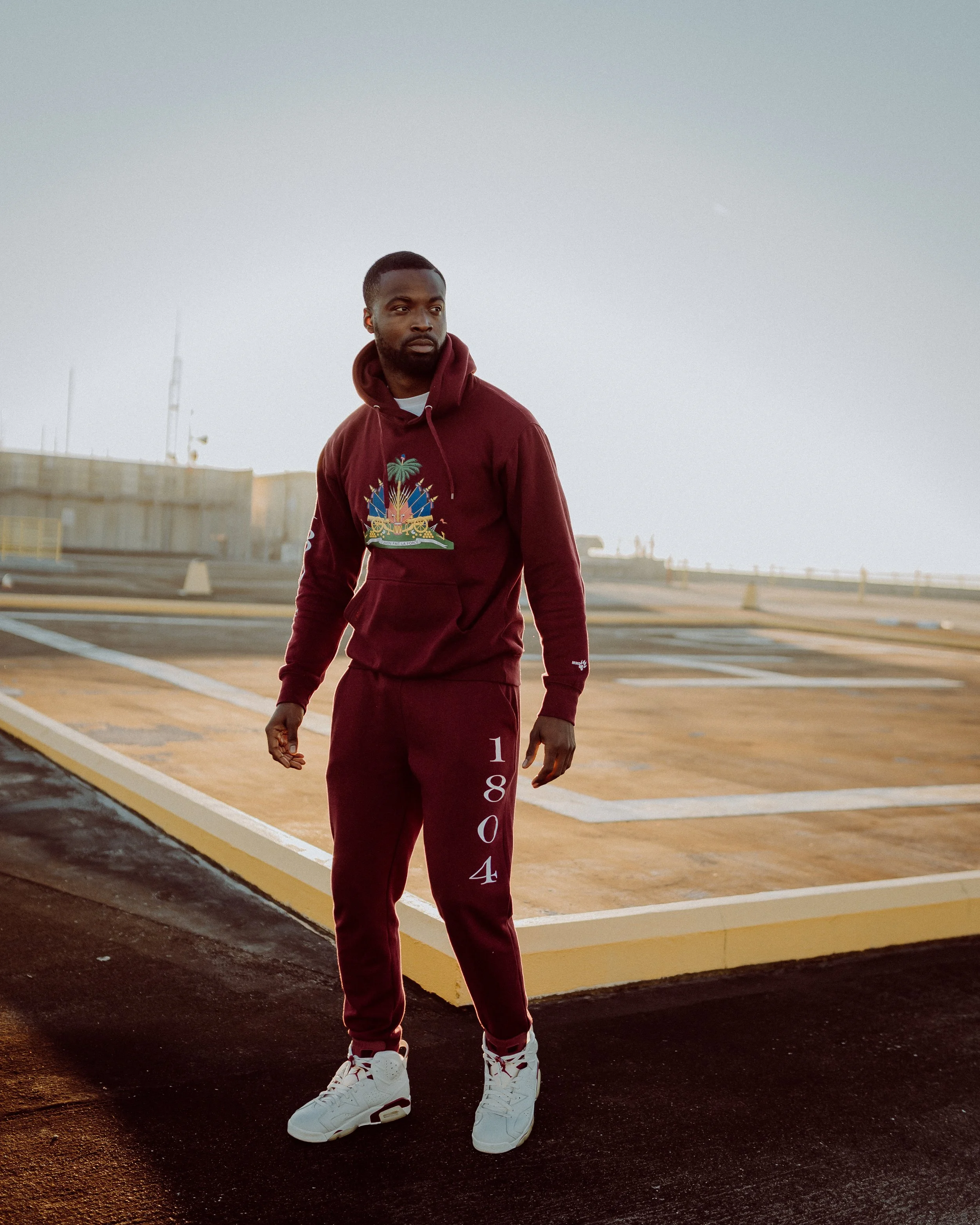 A man in a maroon hoodie and joggers standing on a rooftop parking lot at sunset.