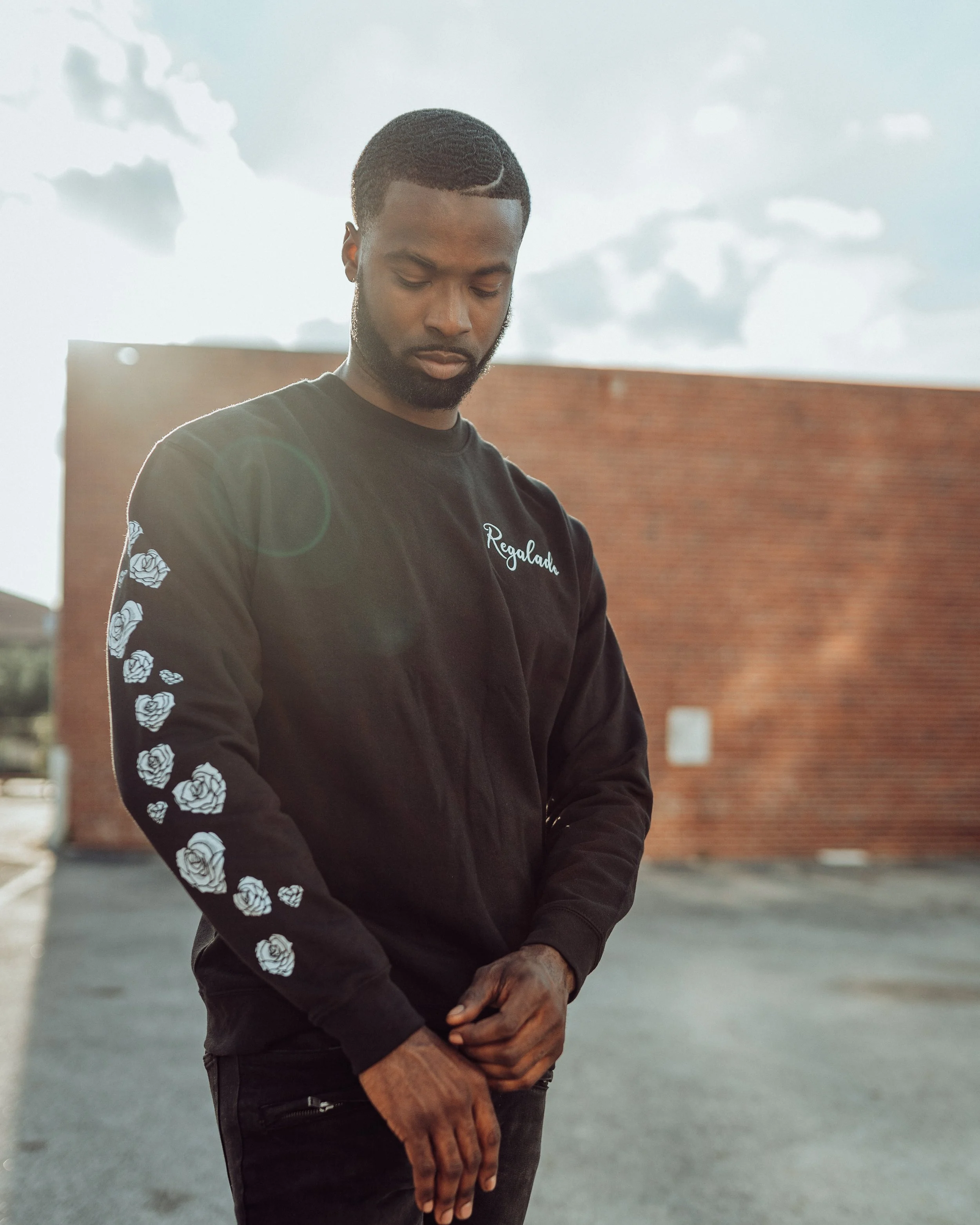 A young man with short curly hair and a beard standing outdoors in front of a brick wall, wearing a black jacket with floral designs on the sleeves and the name "Regalau" embroidered on the chest, looking down with a contemplative expression.