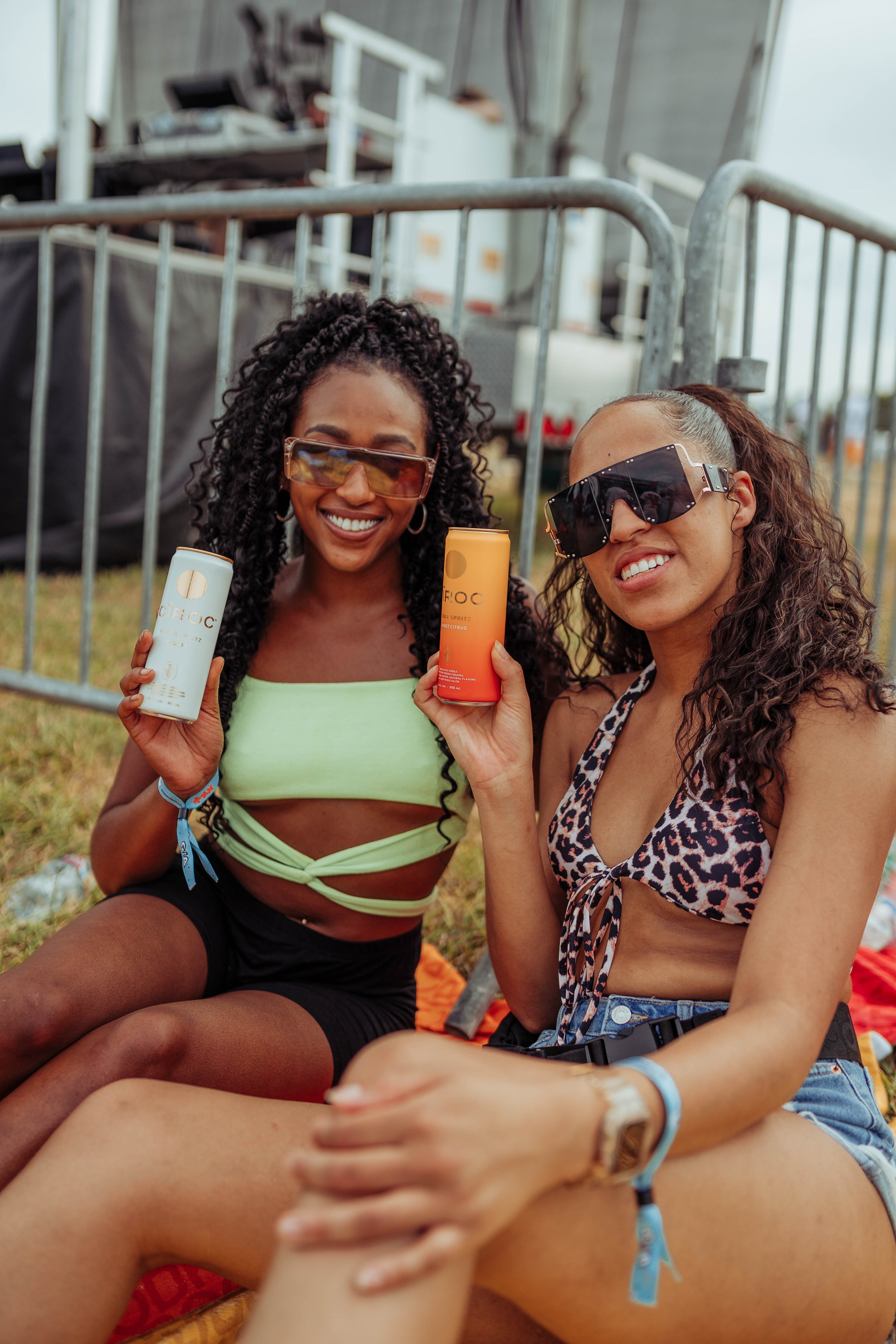 Two women wearing sunglasses and casual summer clothes sitting on the grass at an outdoor event, holding cans of CÎROC vodka drinks, smiling at the camera.