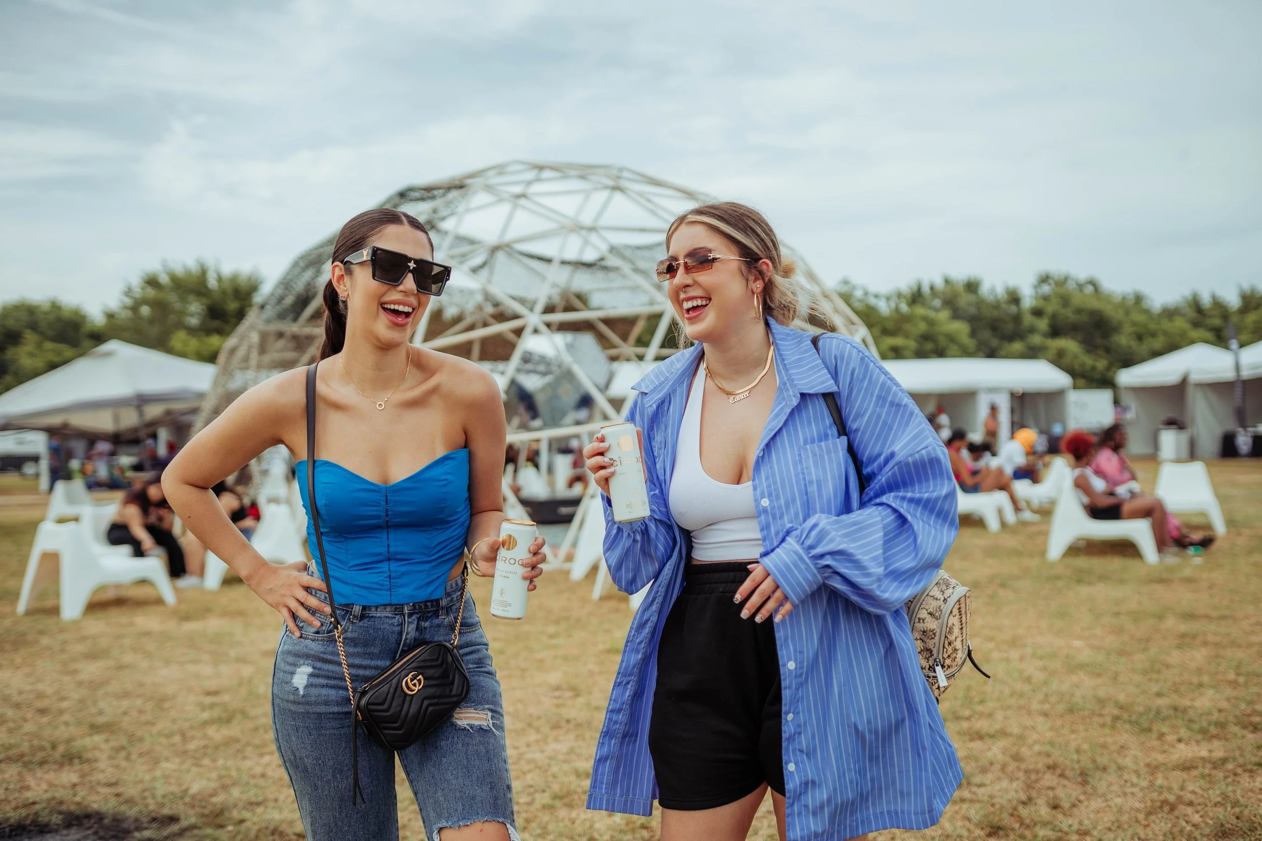 Two women smiling and talking at an outdoor event, dressed casually with sunglasses, holding drinks, with tents and people sitting on white chairs in the background.