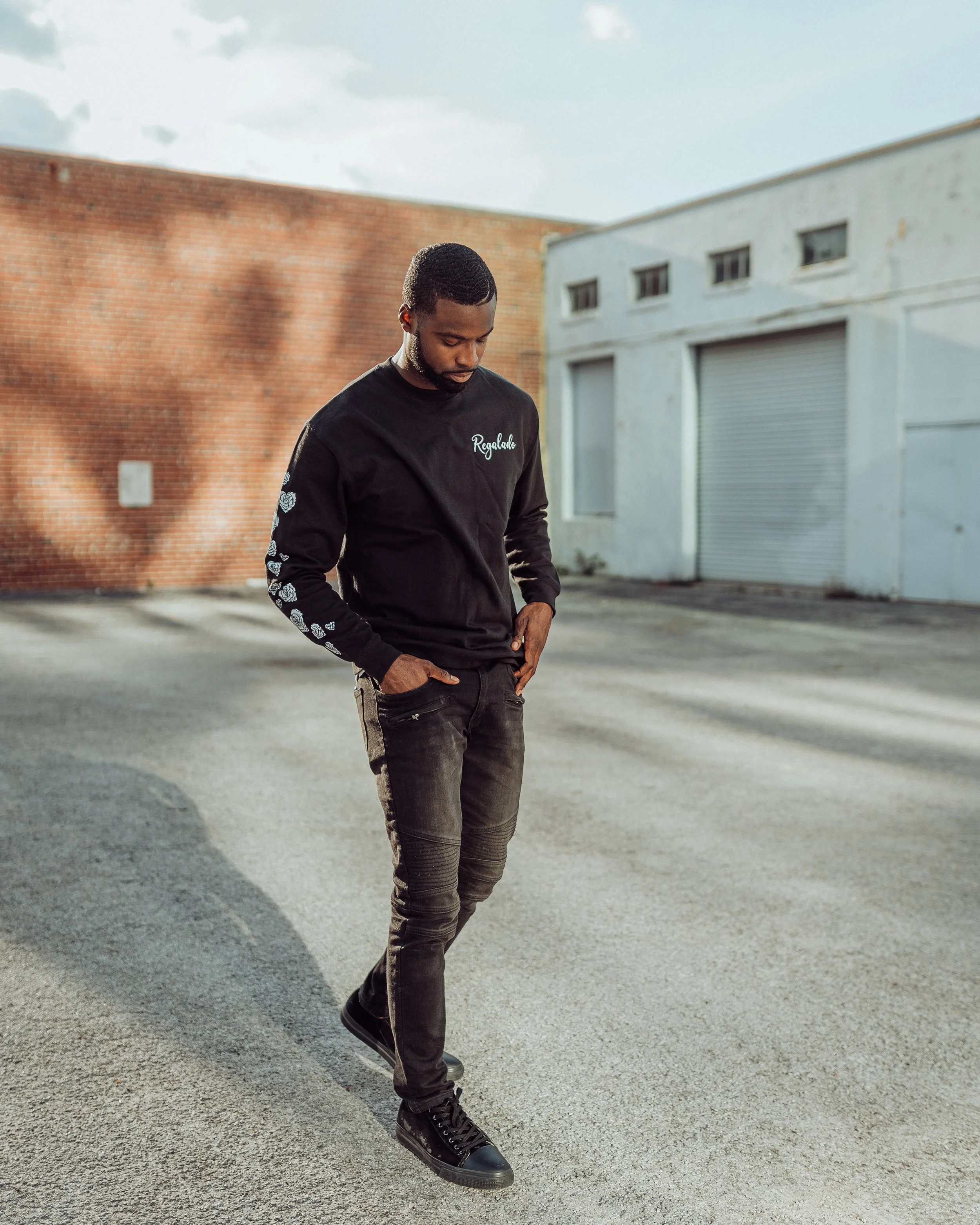 A young man dressed in a black long-sleeve shirt and black pants standing outdoors on a paved street, looking down with hands in pockets, with an industrial background of brick and white buildings.