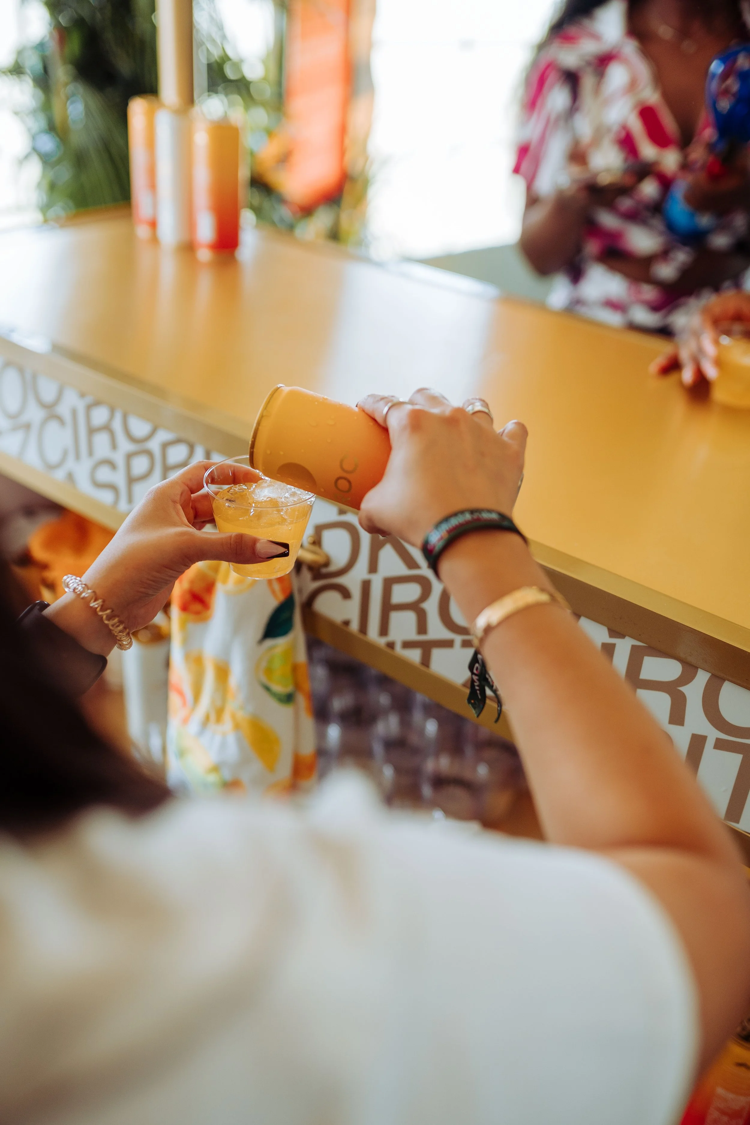 Person pouring a drink from a yellow-orange cup into a small glass, at a colorful bar with orange bottles, with a person in a floral shirt in the background.