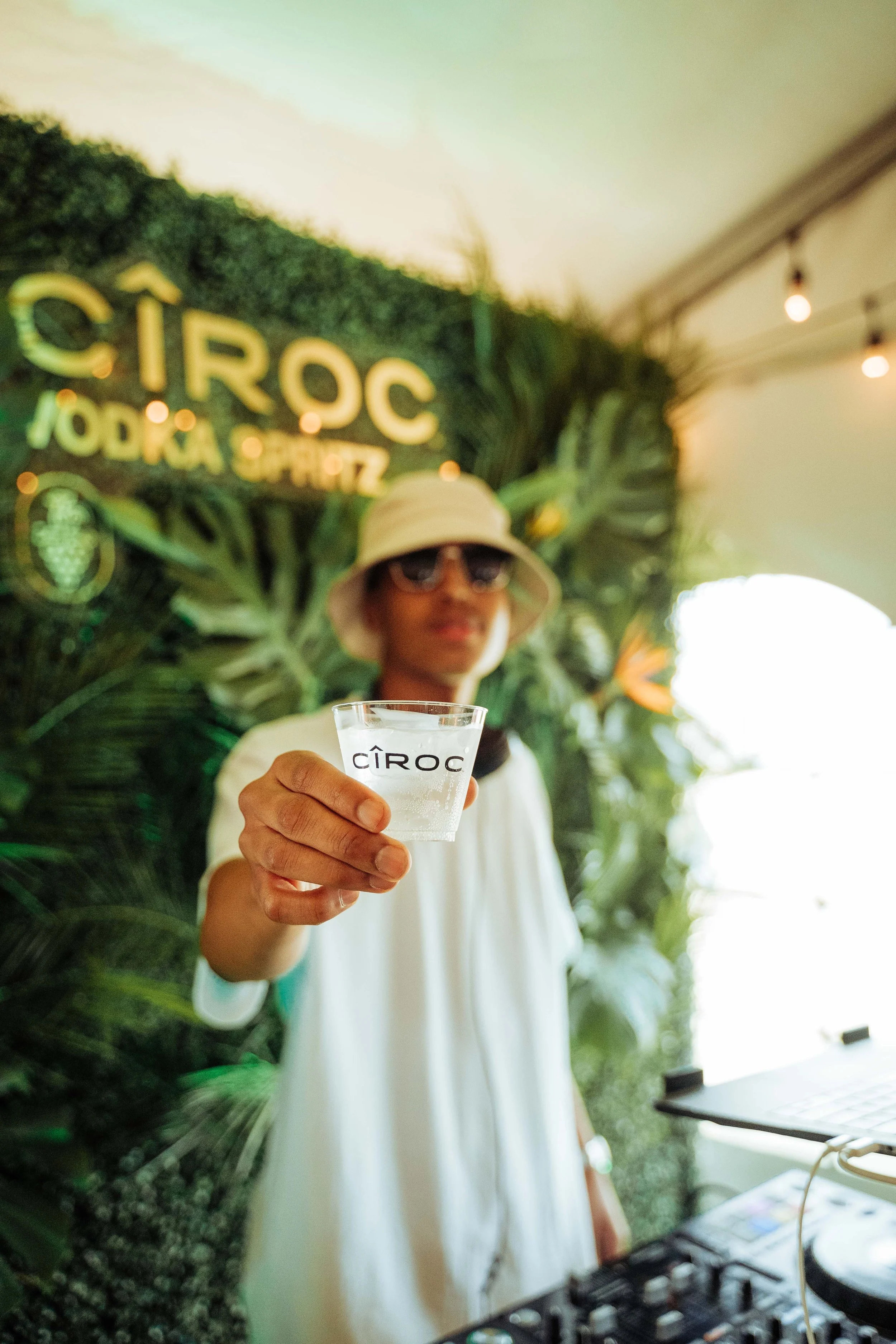 A person in a white outfit and hat holding a clear glass with the CÎROC logo, with a blurred green tropical background and a sign that says 'CÎROC Vodka Spritz' in the background.