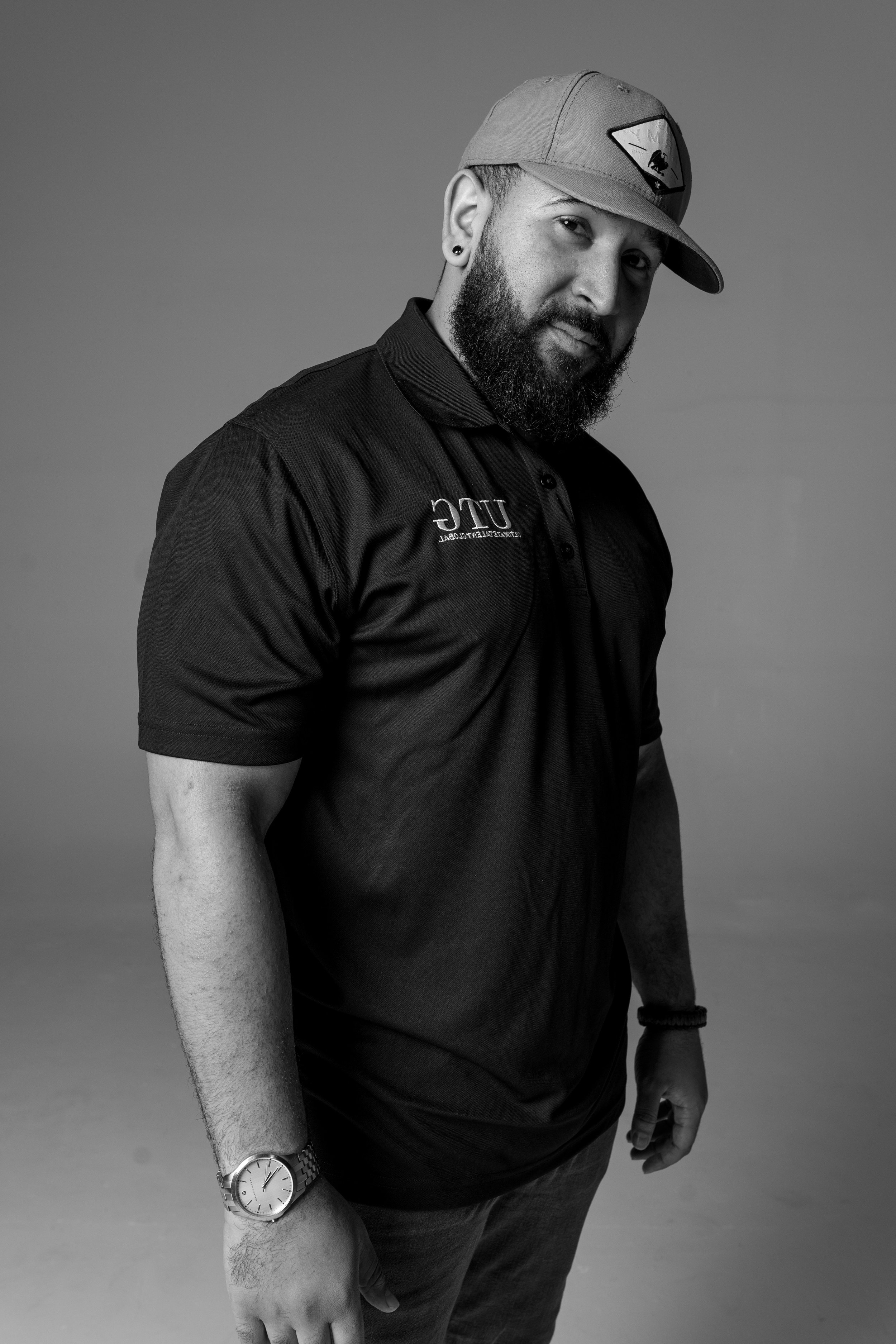 Black and white portrait of a man wearing a cap, a polo shirt, and a watch, standing and looking at the camera.