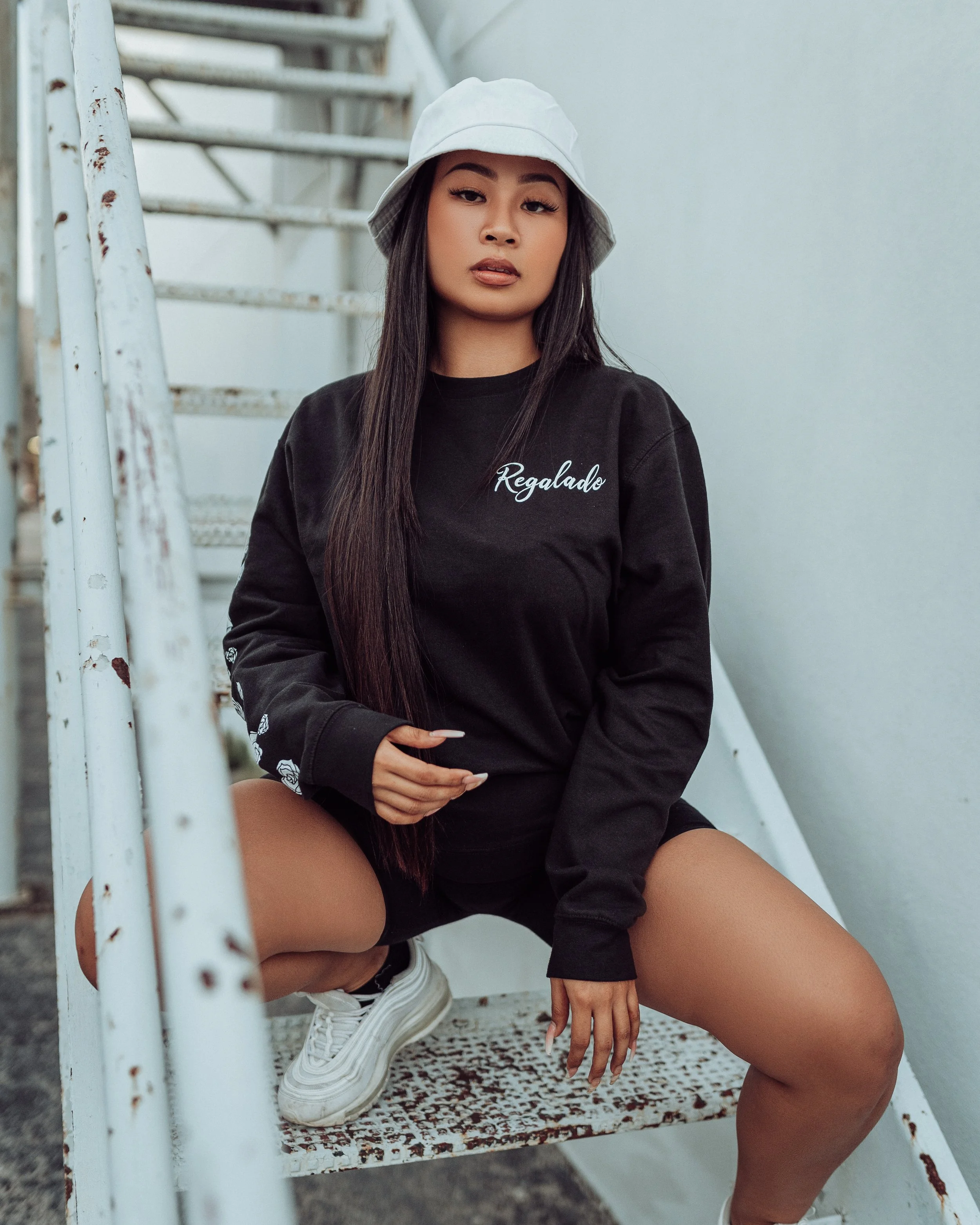 Young woman with long dark hair wearing a white bucket hat, black sweatshirt with white script, black shorts, and white sneakers, is crouching on a rusty outdoor metal staircase.