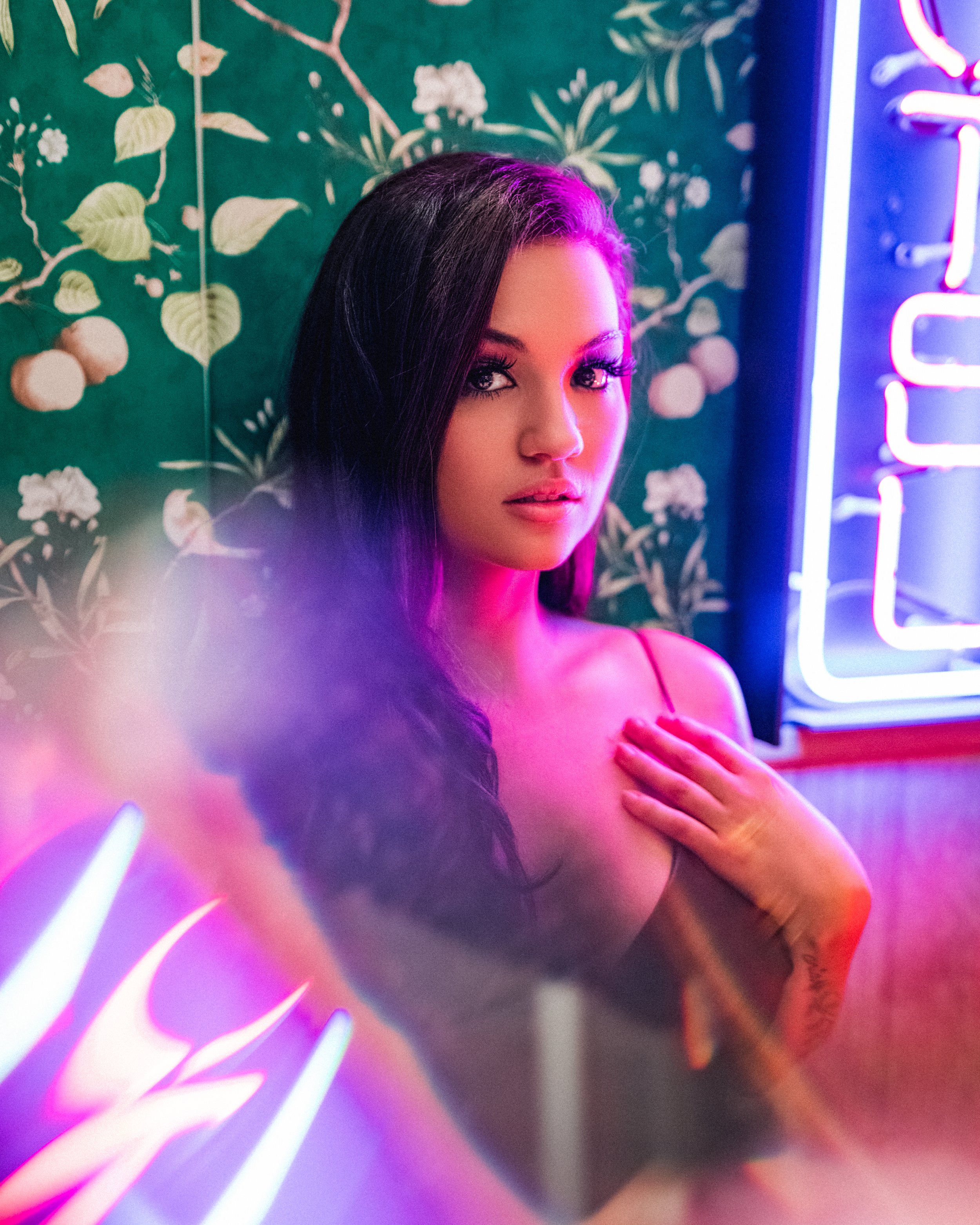 A young woman with long dark hair and makeup, posing in front of a tropical-patterned wallpaper and a neon light, with colorful lighting effects.