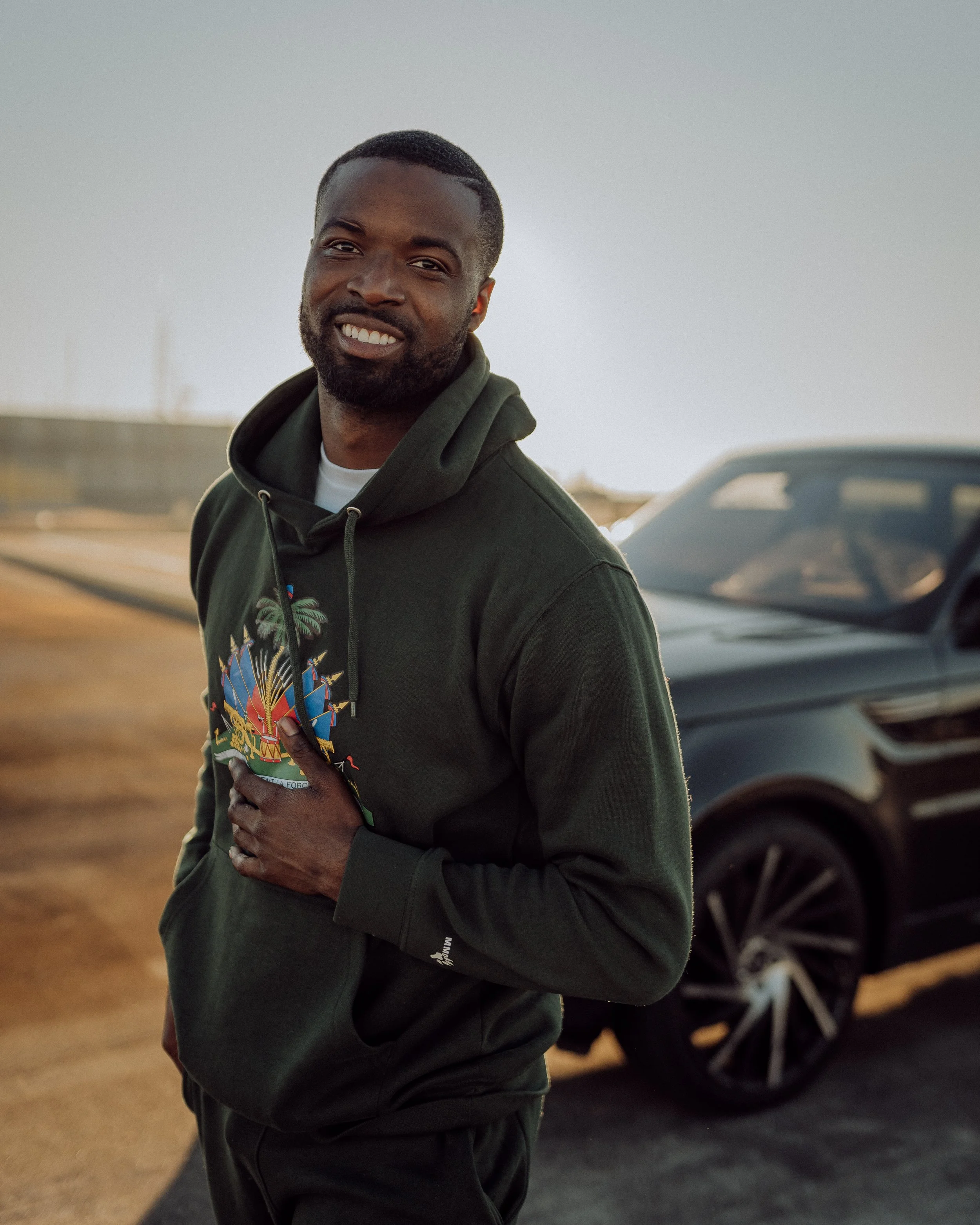 A smiling man standing outdoors near a black car during sunset, wearing a dark green hoodie with colorful graphics.