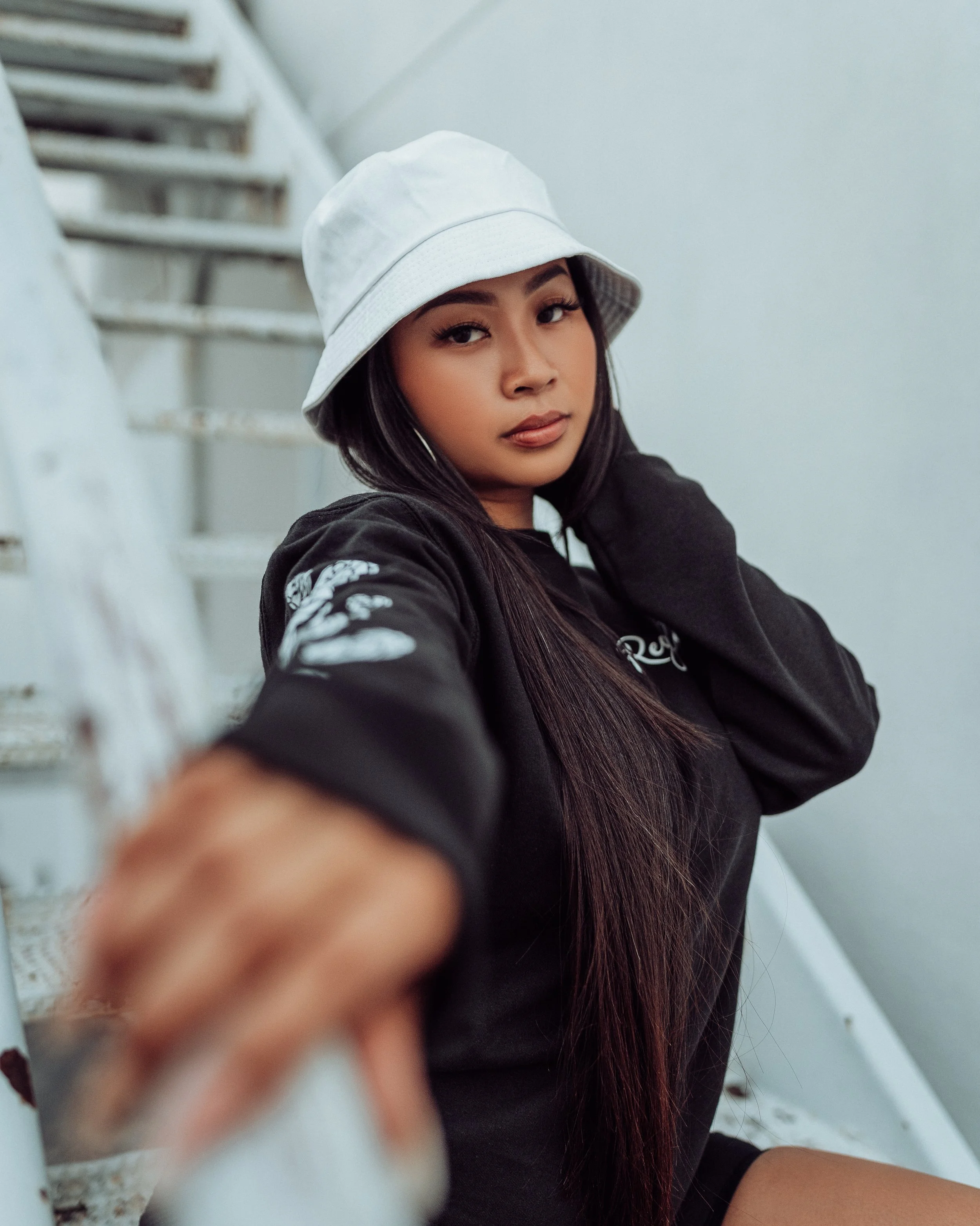 A woman with long dark hair wearing a white bucket hat and a black sweatshirt leaning on a white metal staircase railing, looking at the camera.
