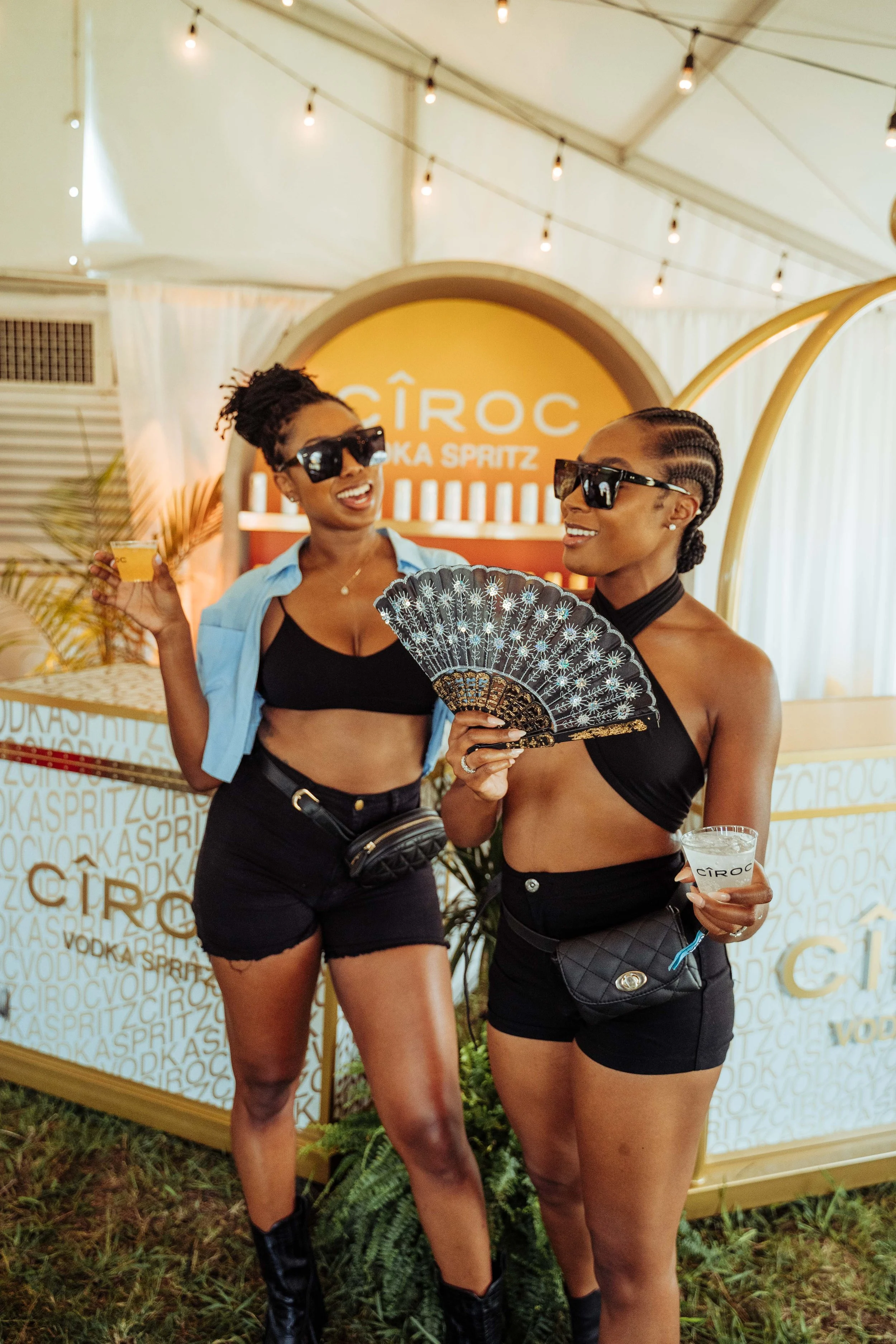 Two women wearing black crop tops and black shorts, with sunglasses, holding drinks and a fan, at a CÎROC vodka event under a white tent with string lights.