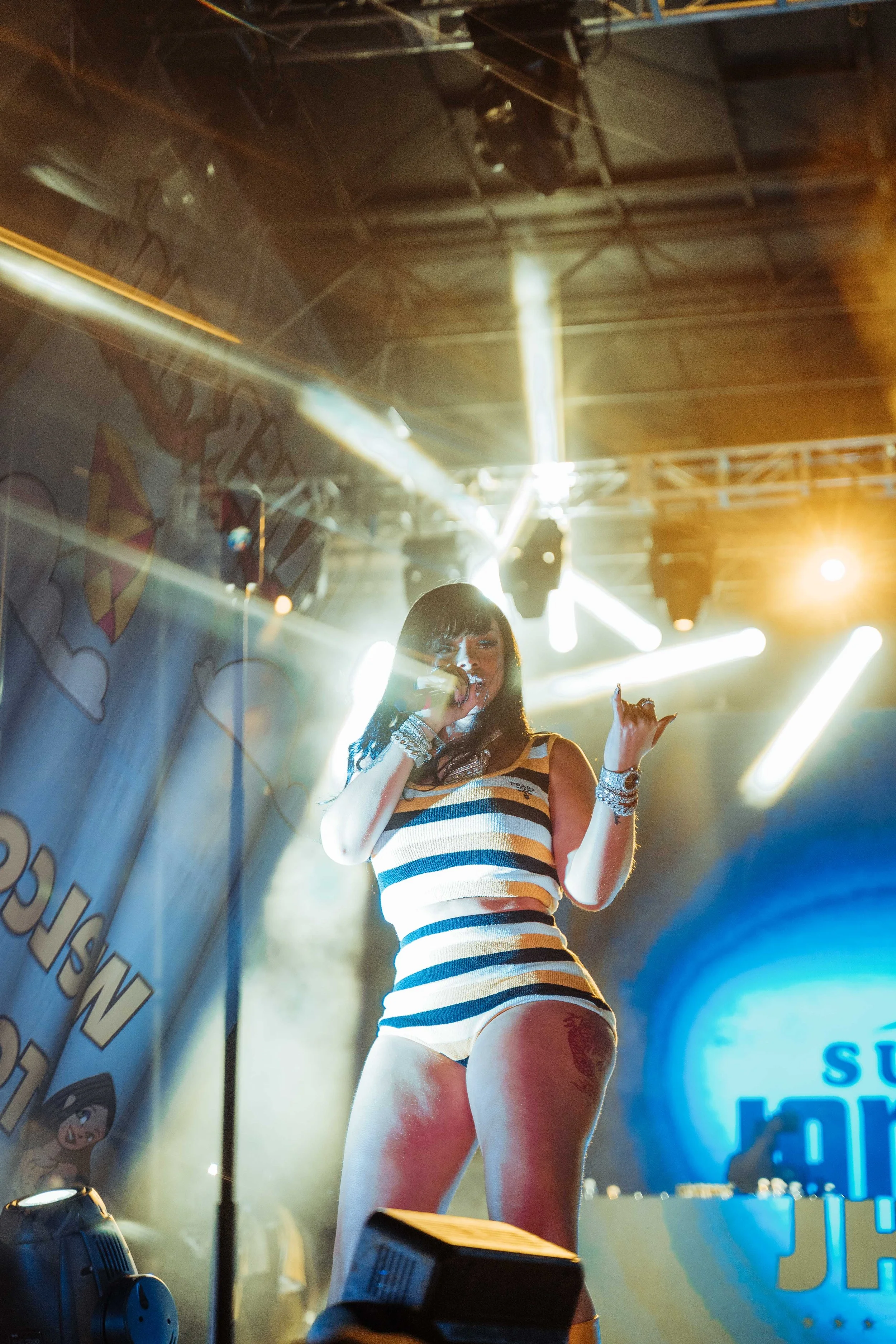 A female performer singing on stage, wearing a striped tank top and shorts, with bright stage lighting and a DJ in the background.