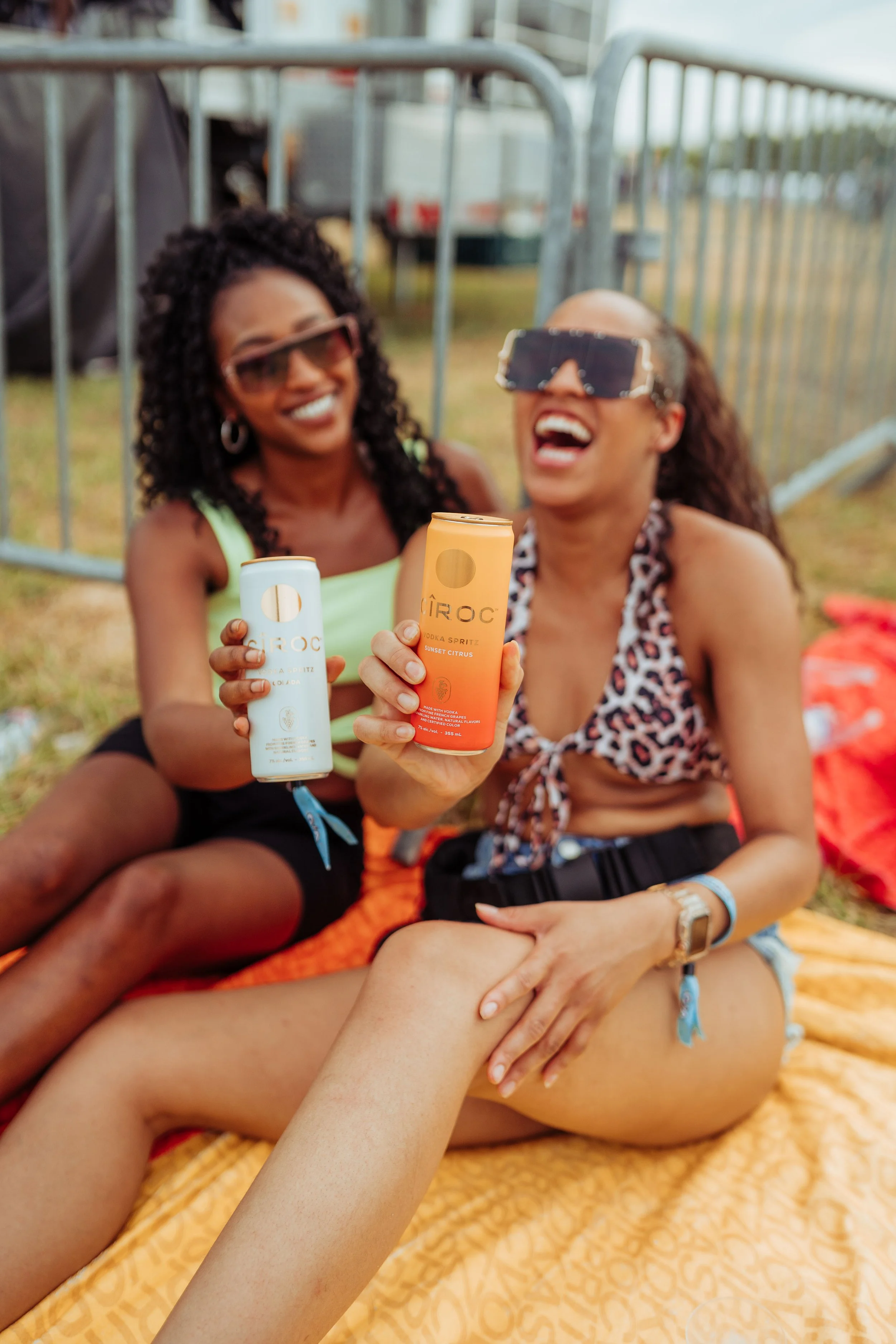 Two women at an outdoor event, smiling and holding canned beverages, sitting on a blanket.