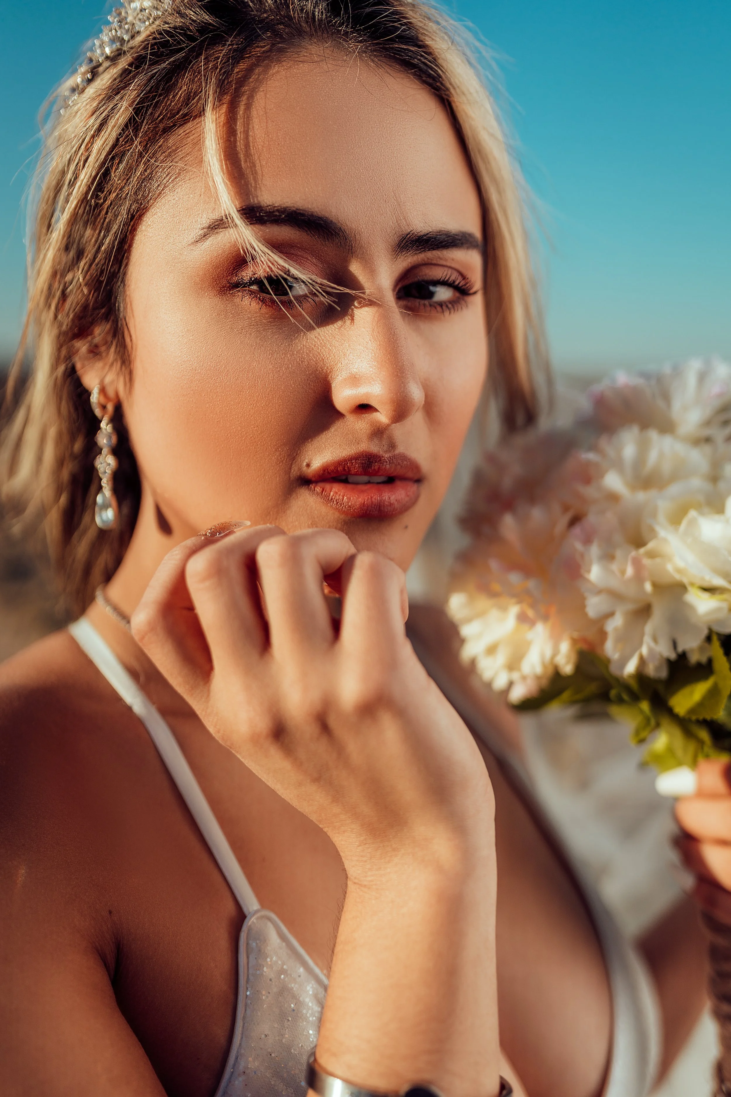 Close-up of a young woman with blonde hair holding a bouquet of white flowers, wearing earrings and a white dress, outdoors on a clear sunny day.