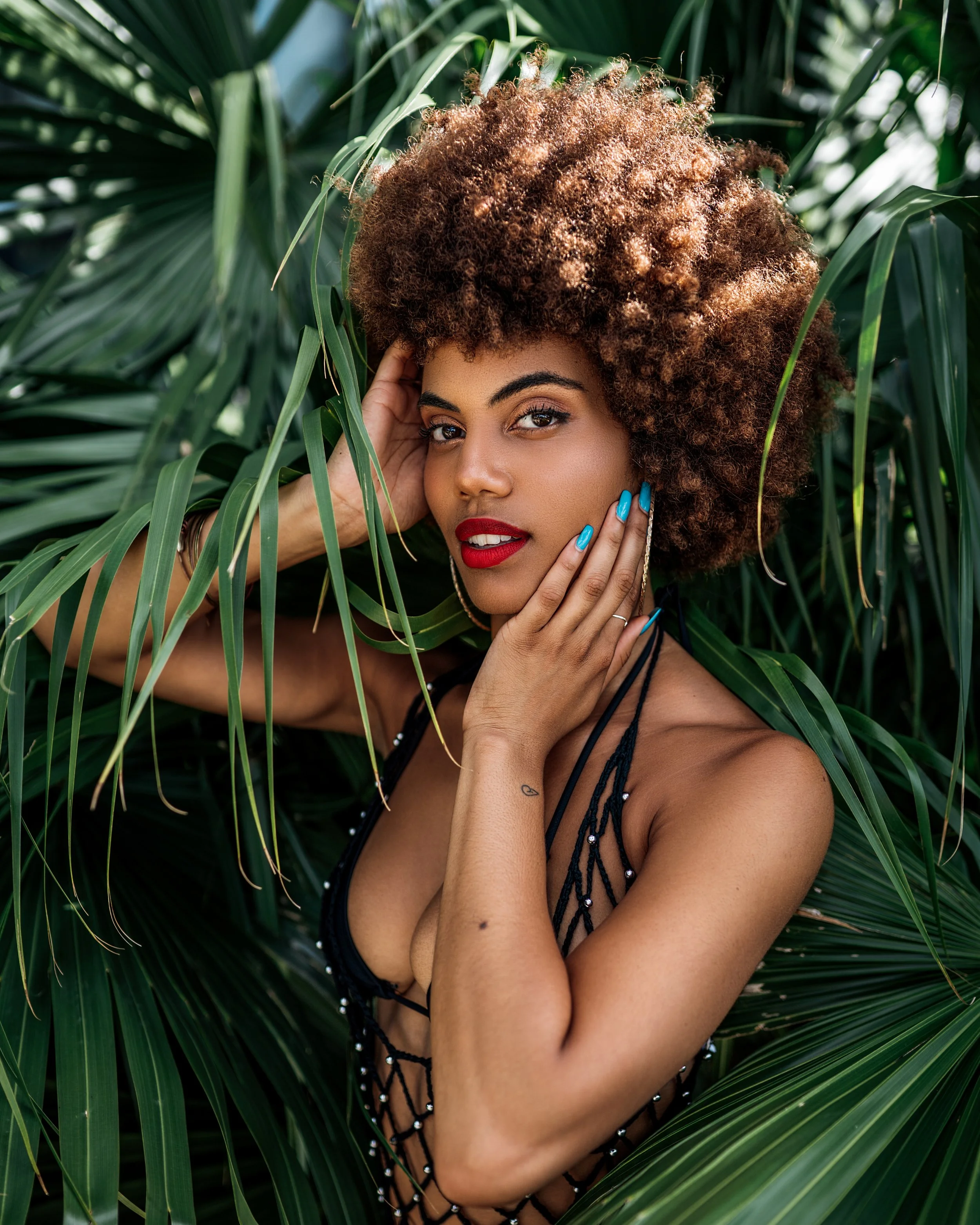 A woman with curly brown hair, red lipstick, and bright blue painted nails posing among green tropical leaves.