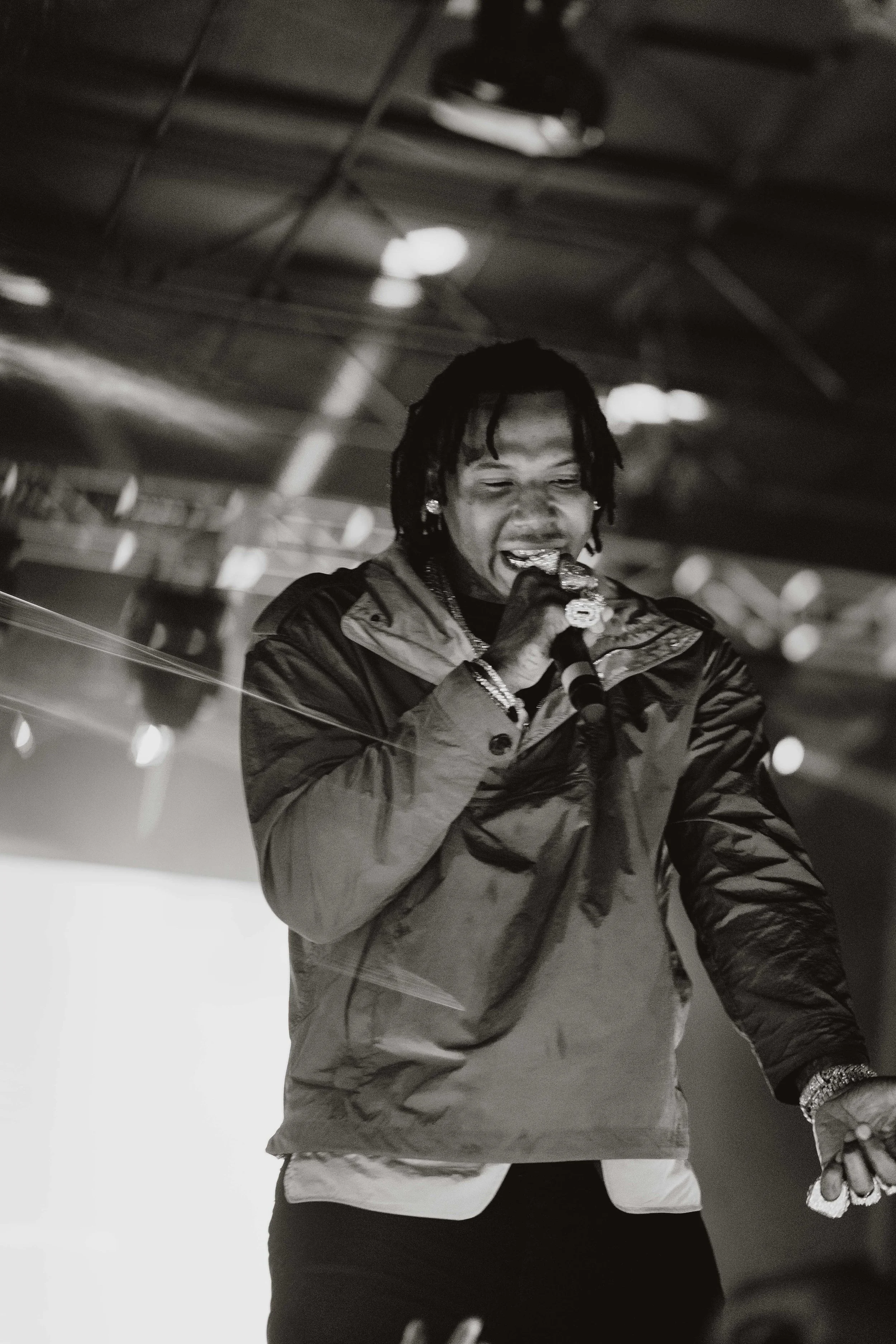 A black and white photo of a man with dreadlocks singing into a microphone on stage, wearing a jacket with an indoor industrial-style ceiling in the background.