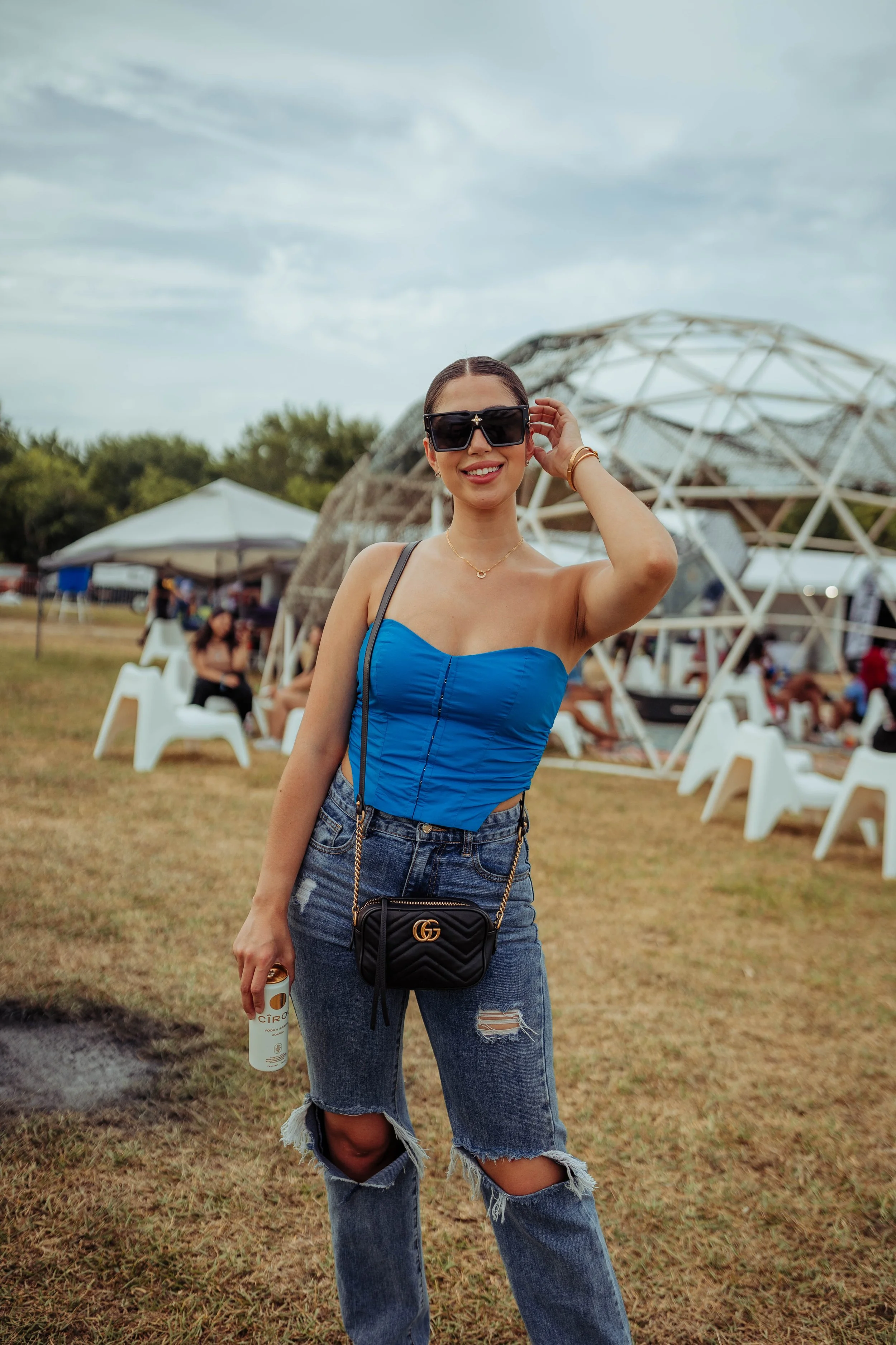 Smiling woman wearing sunglasses, blue top, and ripped jeans, standing outdoors at a festival with geometric stage structure in background.