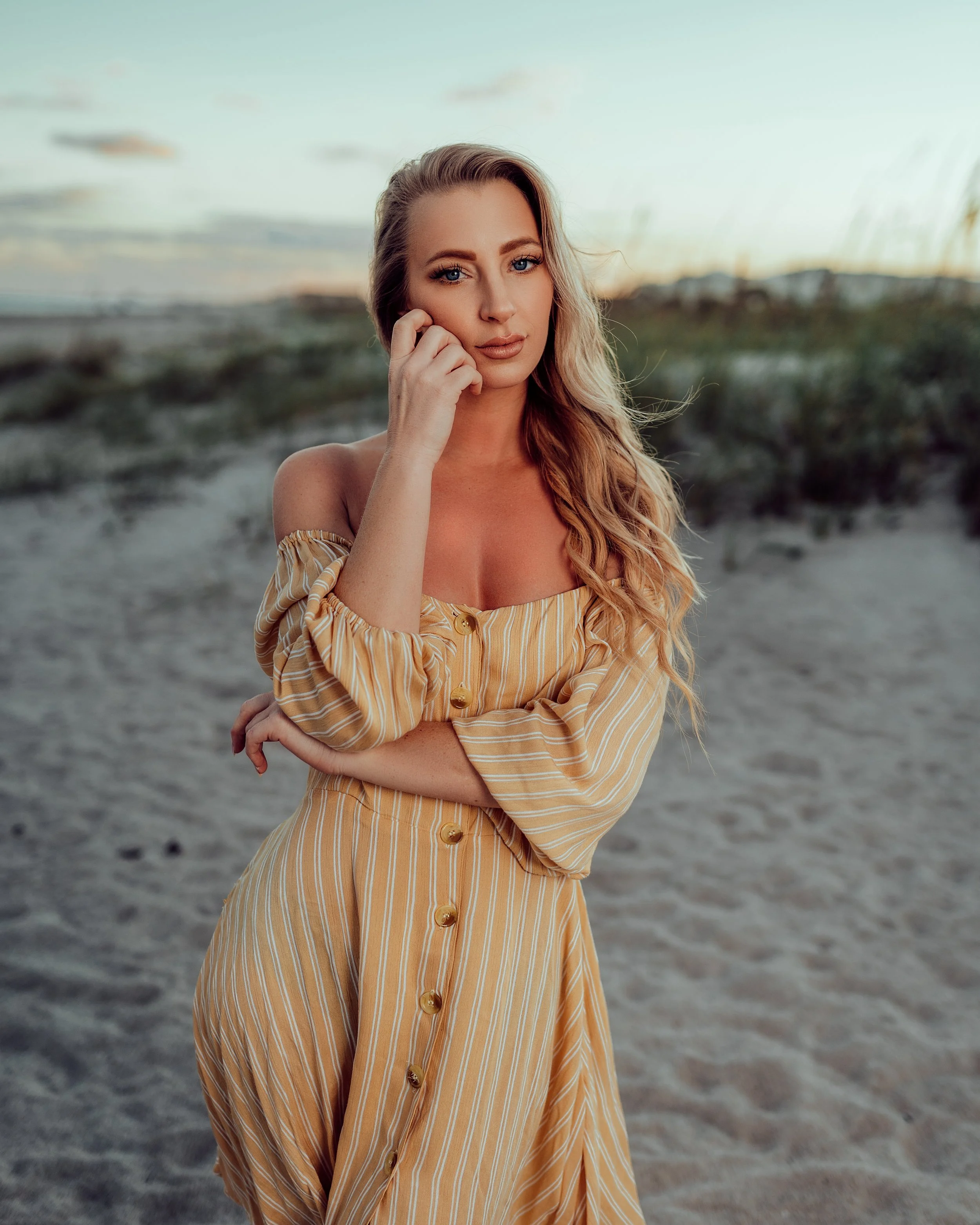 A woman in a yellow and white striped off-the-shoulder dress standing on a sandy beach with dunes in the background, during sunset.
