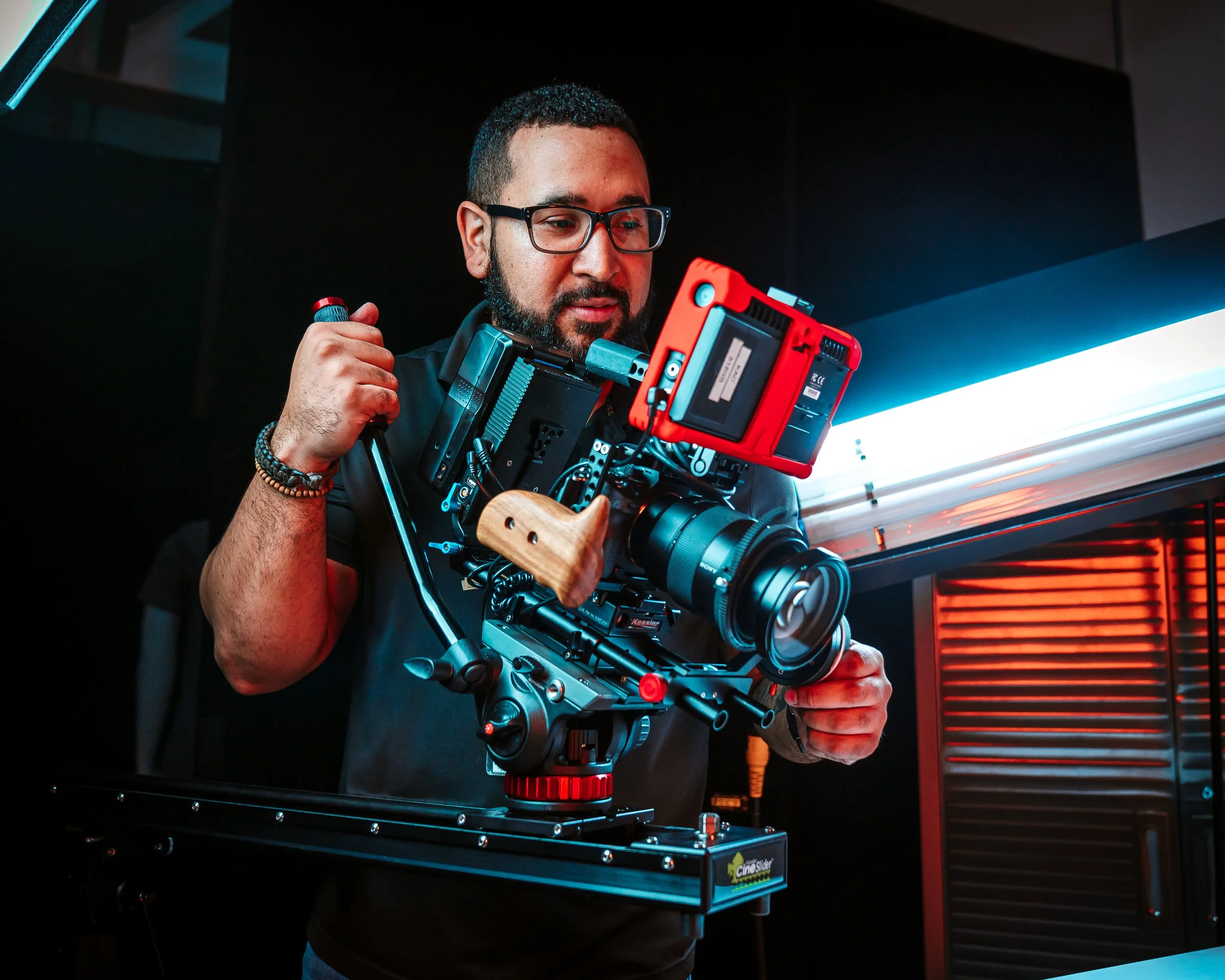 A man operating a professional camera rig with red and black components, in a studio or film set environment with dark walls and neon lighting.