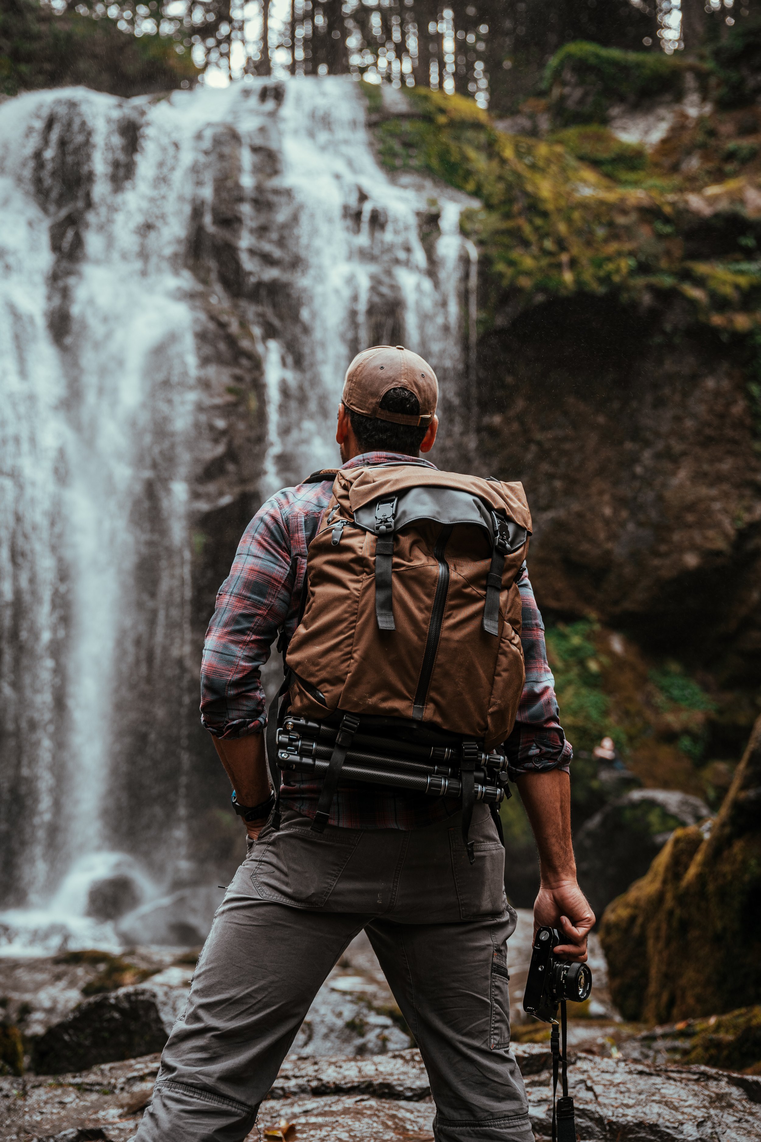 Man with a backpack holding camera stands in front of a waterfall in a forest