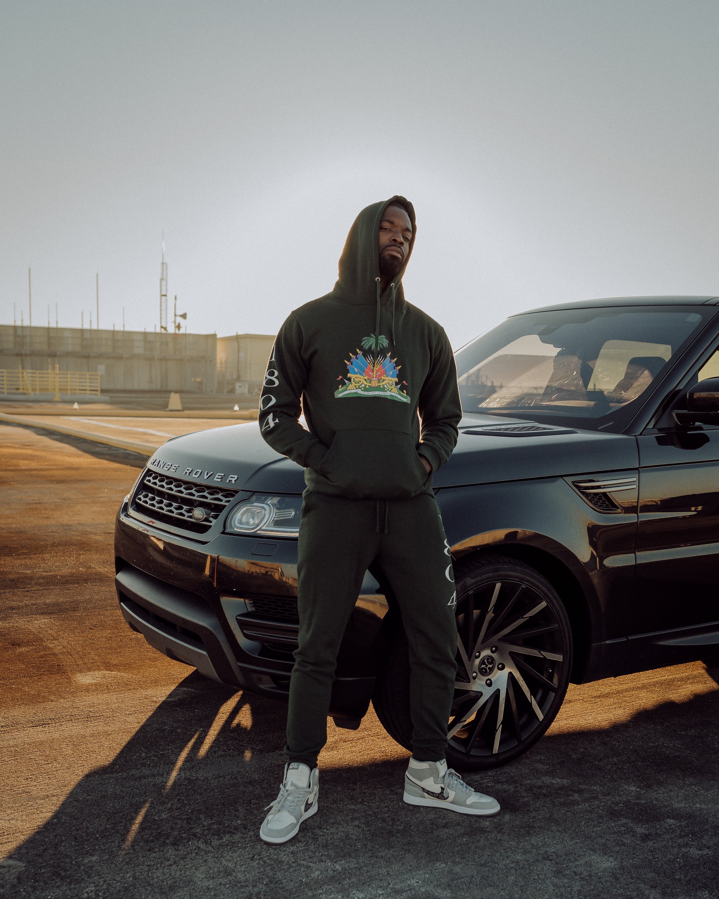 A man in a black hoodie and sweatpants stands with hands in pockets in front of a black Range Rover on a rooftop parking lot during sunset.