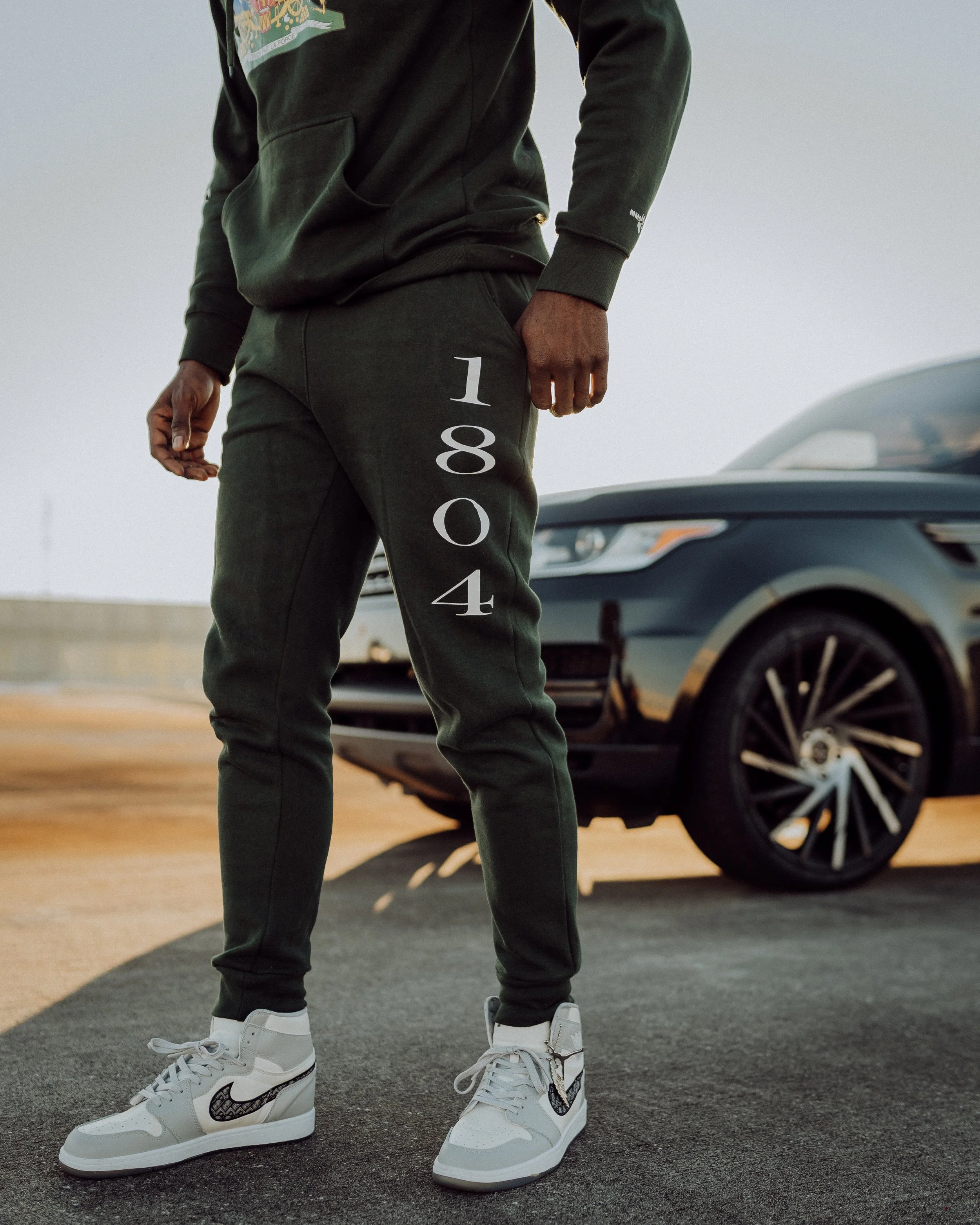 Person wearing dark green hoodie and sweatpants with '1804' printed on the pants, standing outdoors next to a black car, with white sneakers.