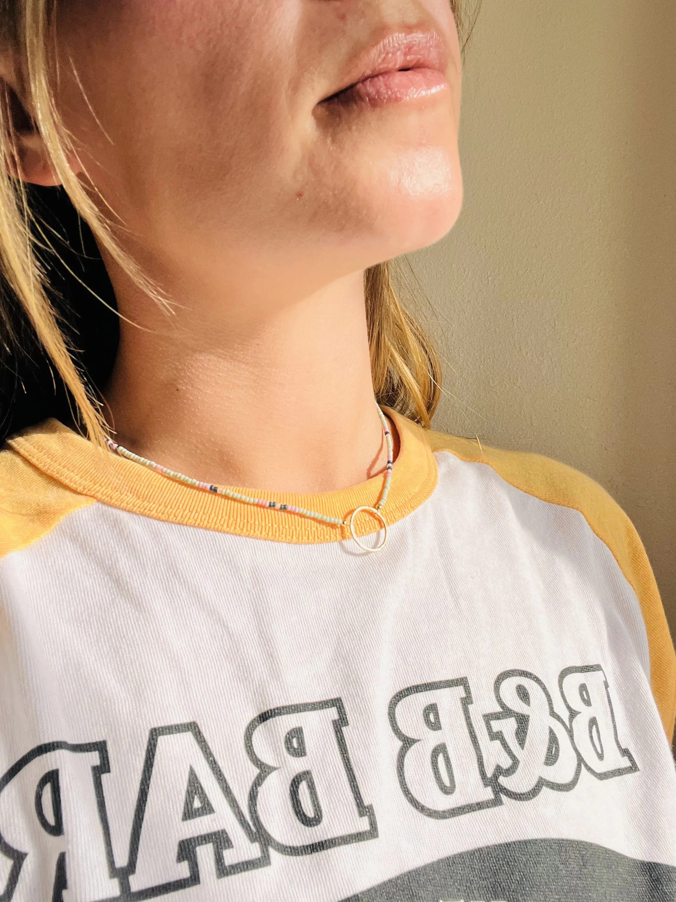 Close-up of a woman’s lower face and neck wearing a yellow and white shirt, a colorful beaded necklace, and a silver ring choker.