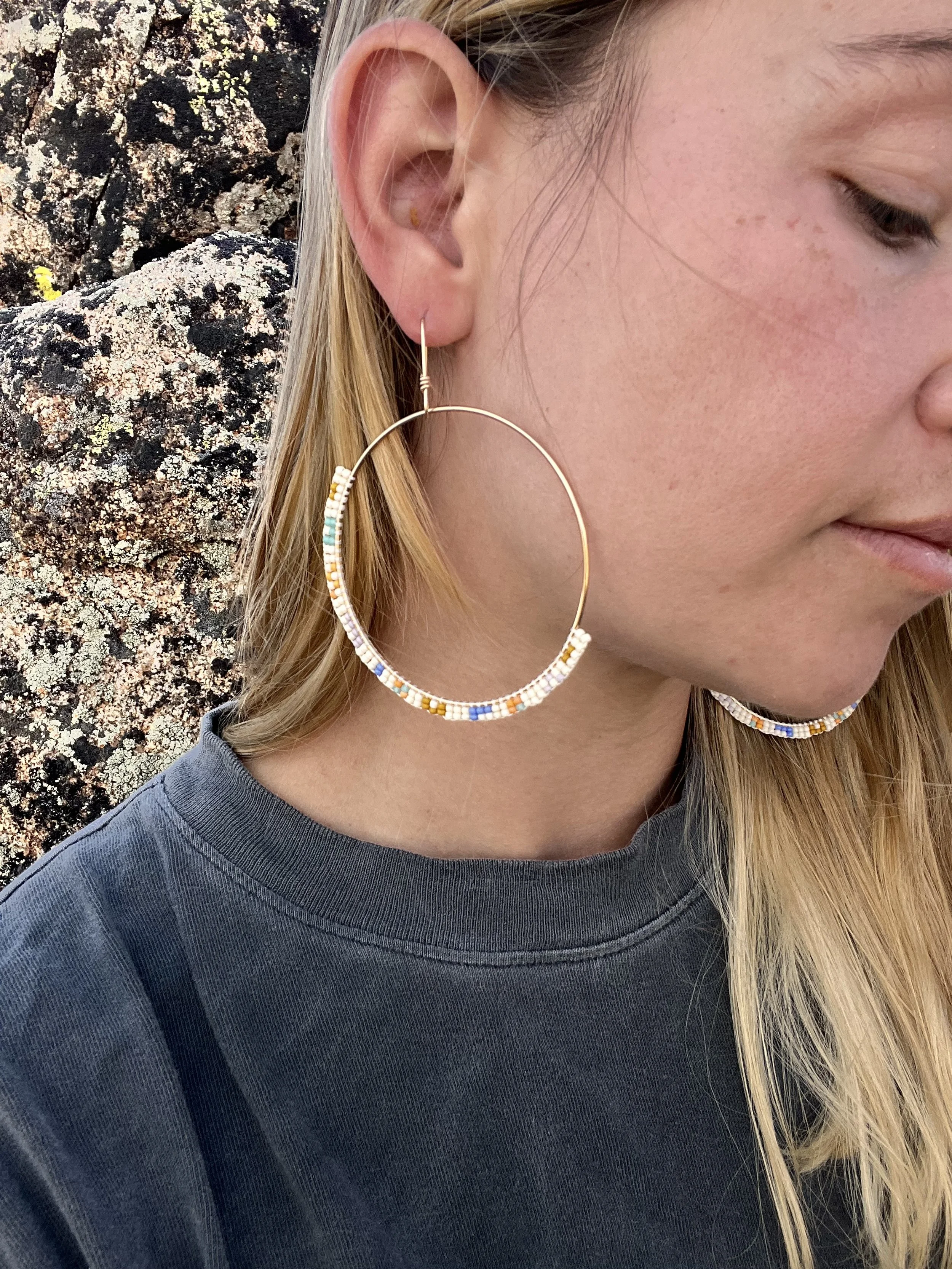 Close-up of a woman's face and shoulder showing her wearing a large, beaded hoop earring and a dark gray shirt with a rocky background.