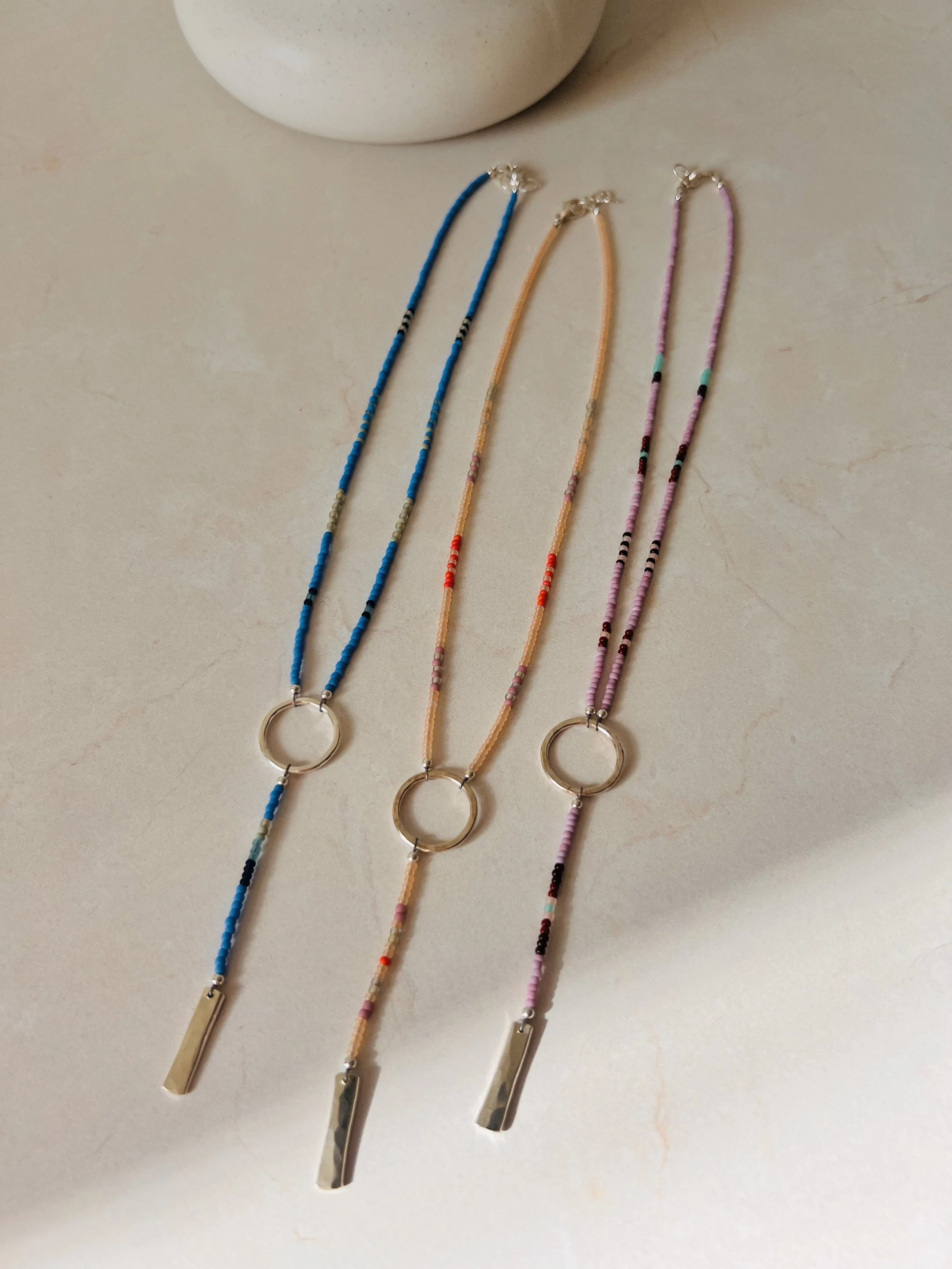 Three beaded necklaces with metallic pendants on a light-colored surface, with a white object partially visible in the background.