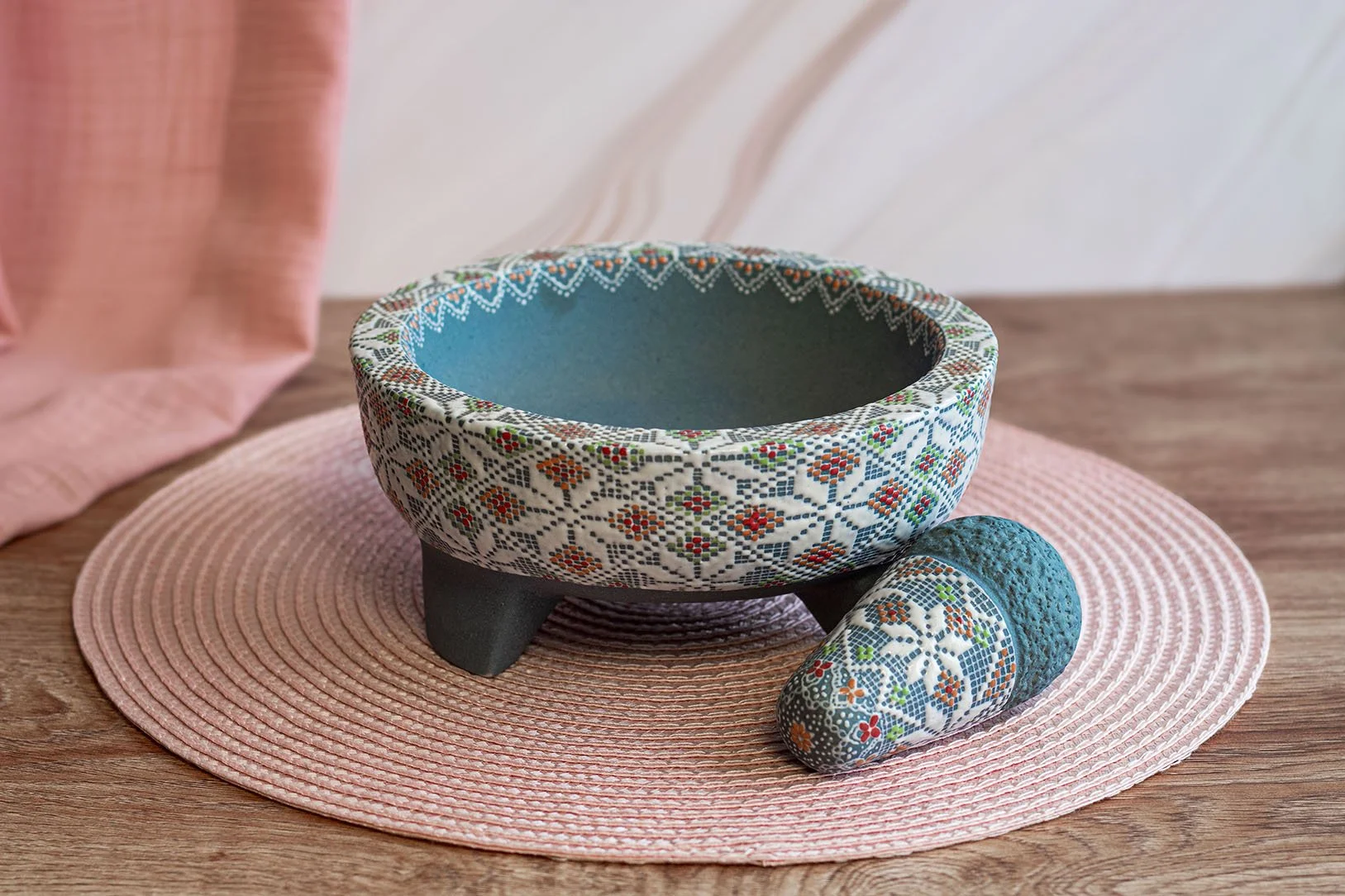 Cuenco decorativo con patrón multicolor en una base negra, acompañado de una piedra decorativa con diseño similar, todo en una base de tela de color rosa sobre mesa de madera.
