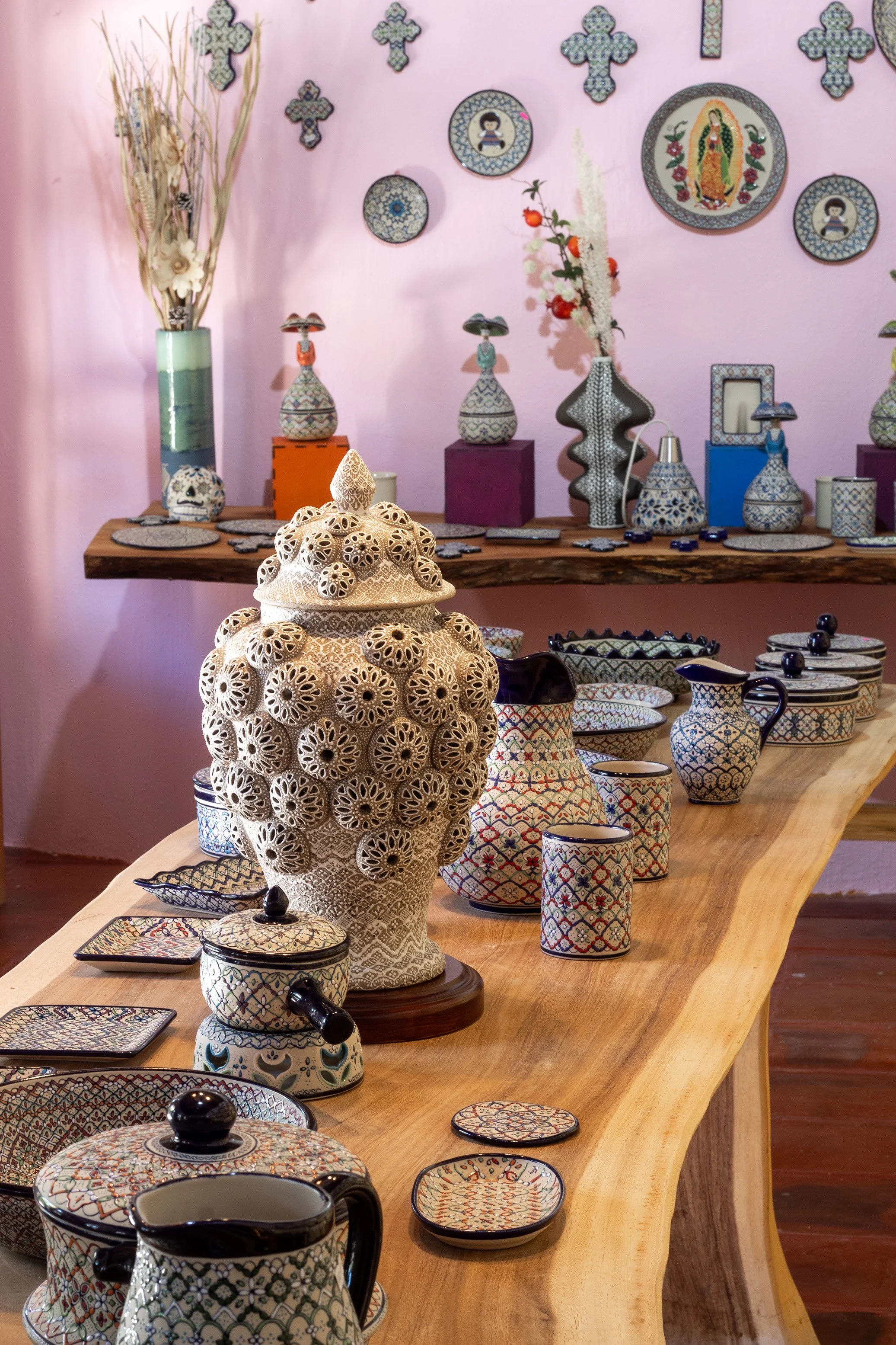 Varios artículos de cerámica decorativa y luminarias, incluyendo jarrones, vasos, platos y una lámpara grande, en una mesa de madera en una tienda con paredes rosadas y arte popular en el fondo en la tienda de Eva Servin Cerámica