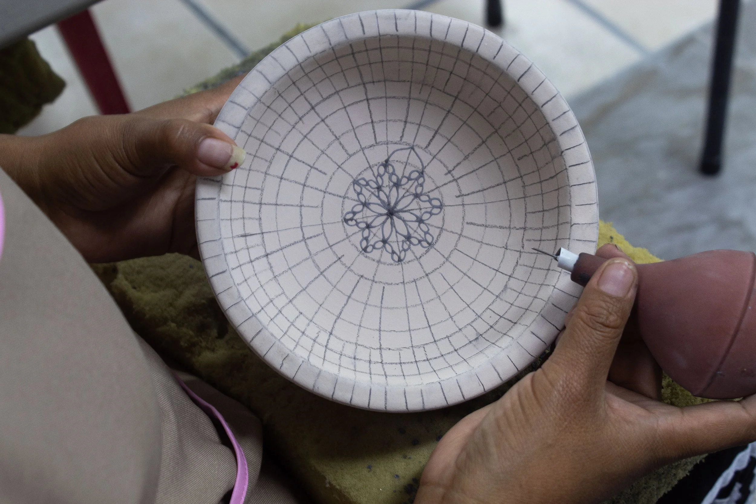 Mano sosteniendo un plato de cerámica con un diseño en el centro y un bolígrafo, en un espacio de trabajo.