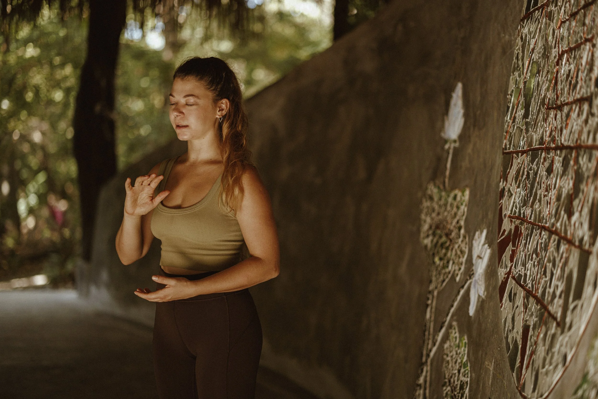 A woman dressed in a tank top and dark pants standing outdoors beside a tree, with her eyes closed and one hand on her chest and the other on her stomach, in a peaceful pose.