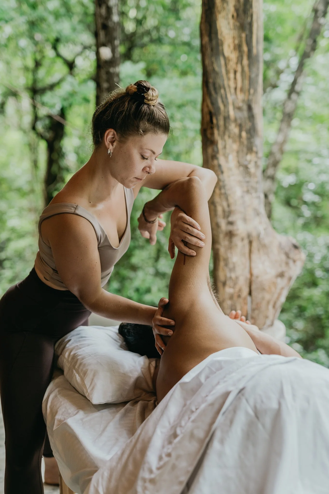 A woman receives a massage in a natural outdoor setting with lush greenery and trees.