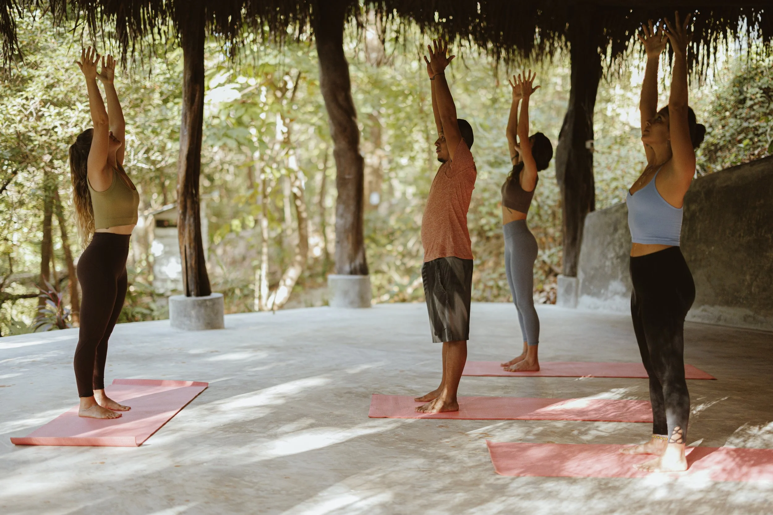 Four people practicing yoga outdoors on pink mats under a thatched roof, standing in a pose with arms raised overhead, surrounded by trees and natural scenery.