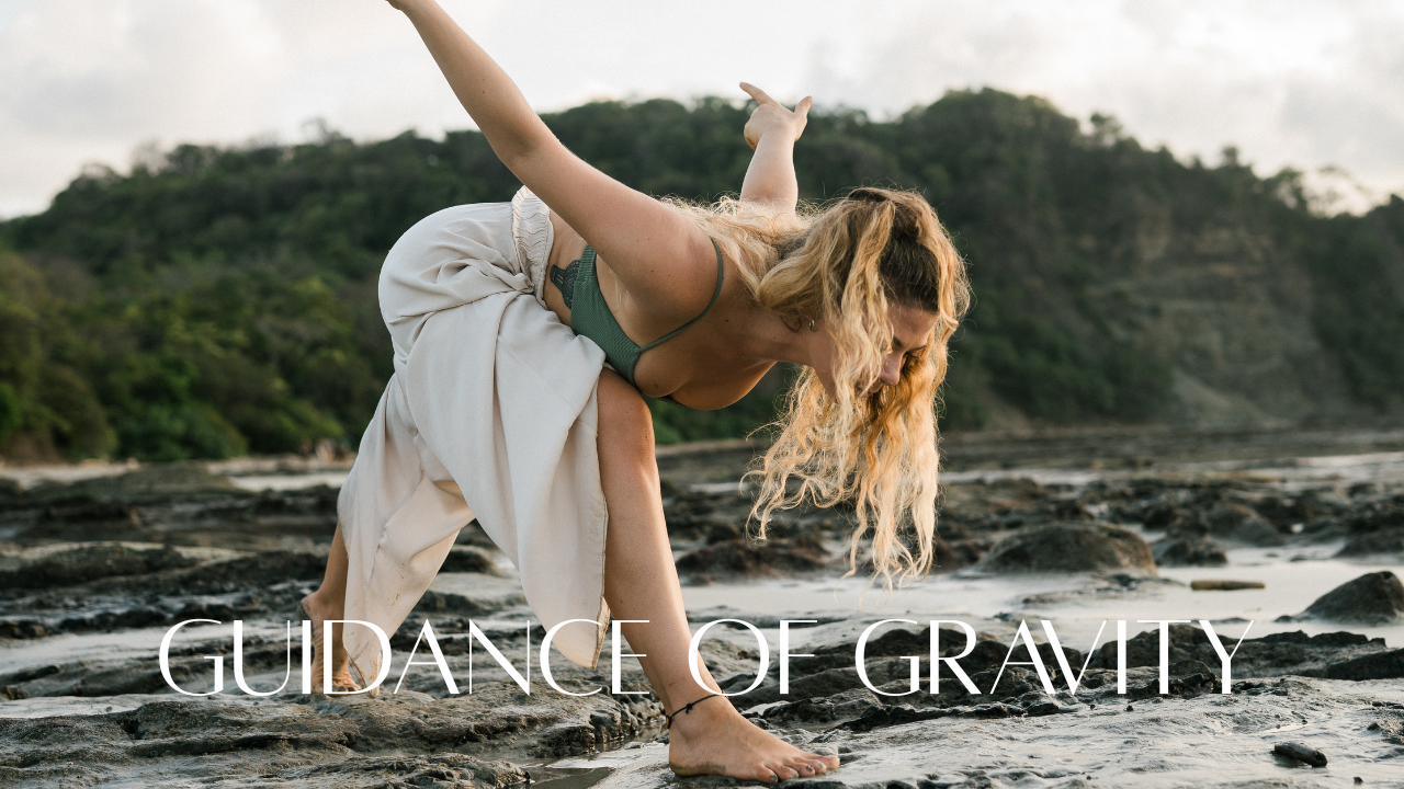 Woman practicing yoga on rocky beach near ocean with a backdrop of green hills, bending over with one hand on the ground and the other arm raised, with text "Guidance of Gravity" overlaid.