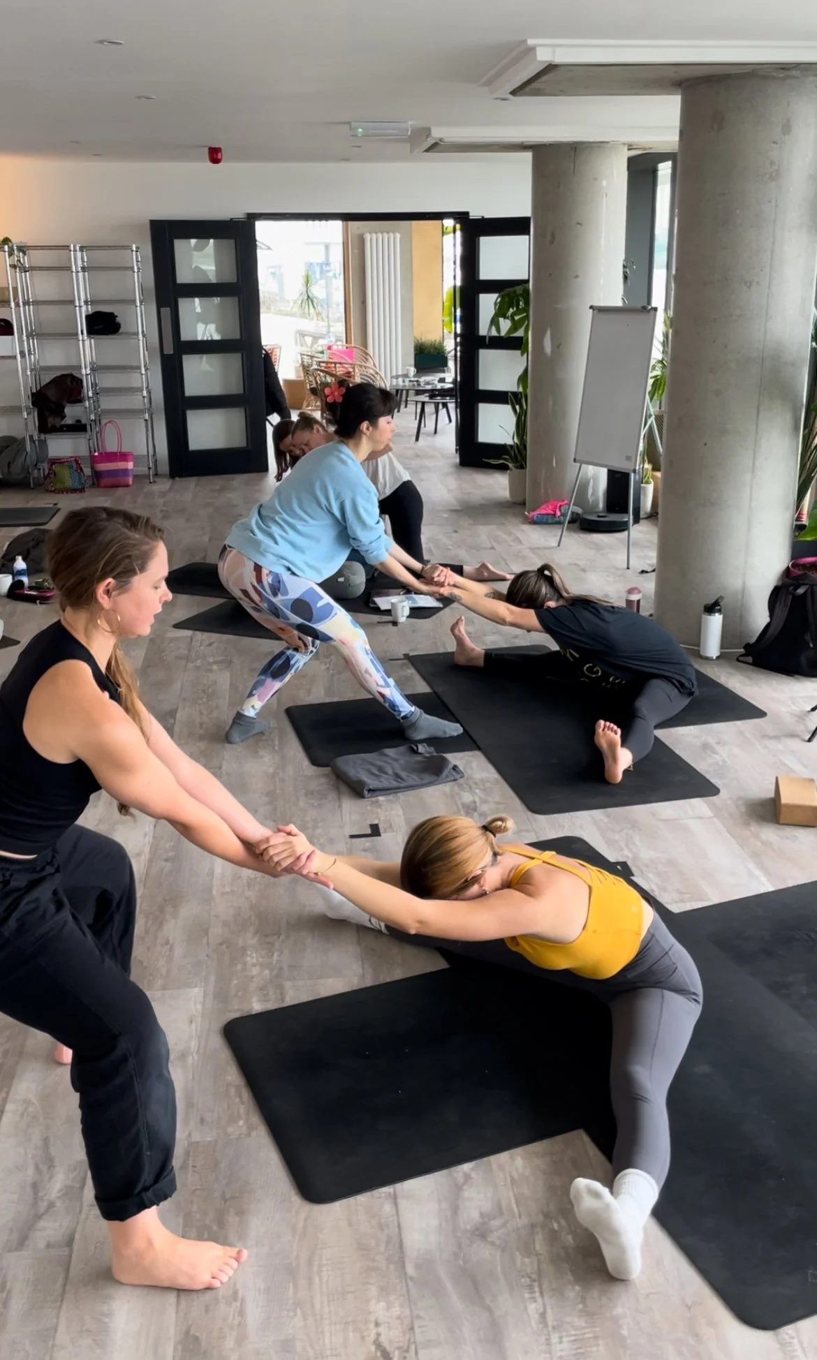 Yoga class with woman in yellow top and gray leggings doing a reclining pose, assisted by instructor in black pants, with other participants in background performing partner yoga stretches.