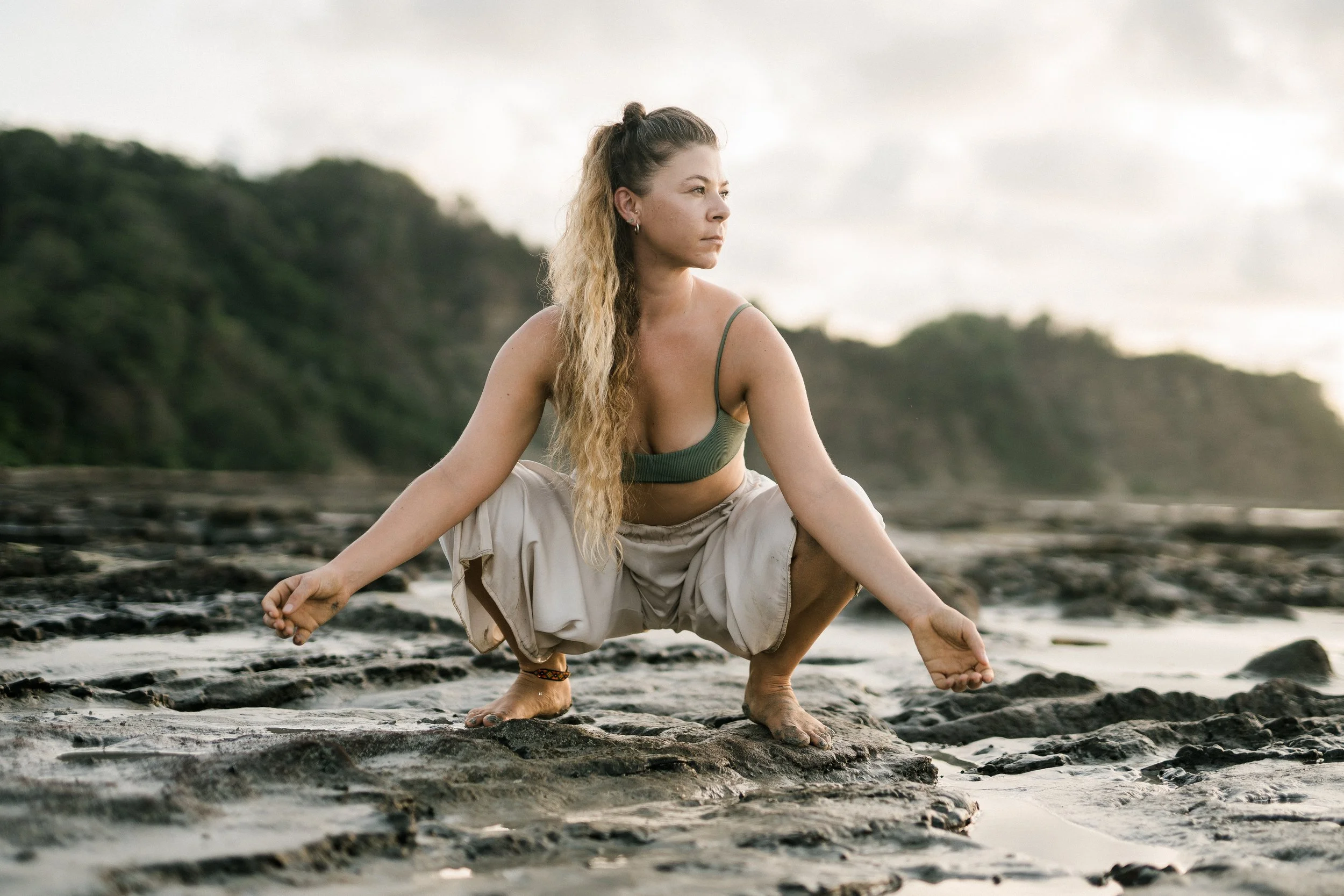 Woman practicing yoga on rocky beach at sunset or sunrise, squatting with hands on ground, in green sports bra and beige pants.