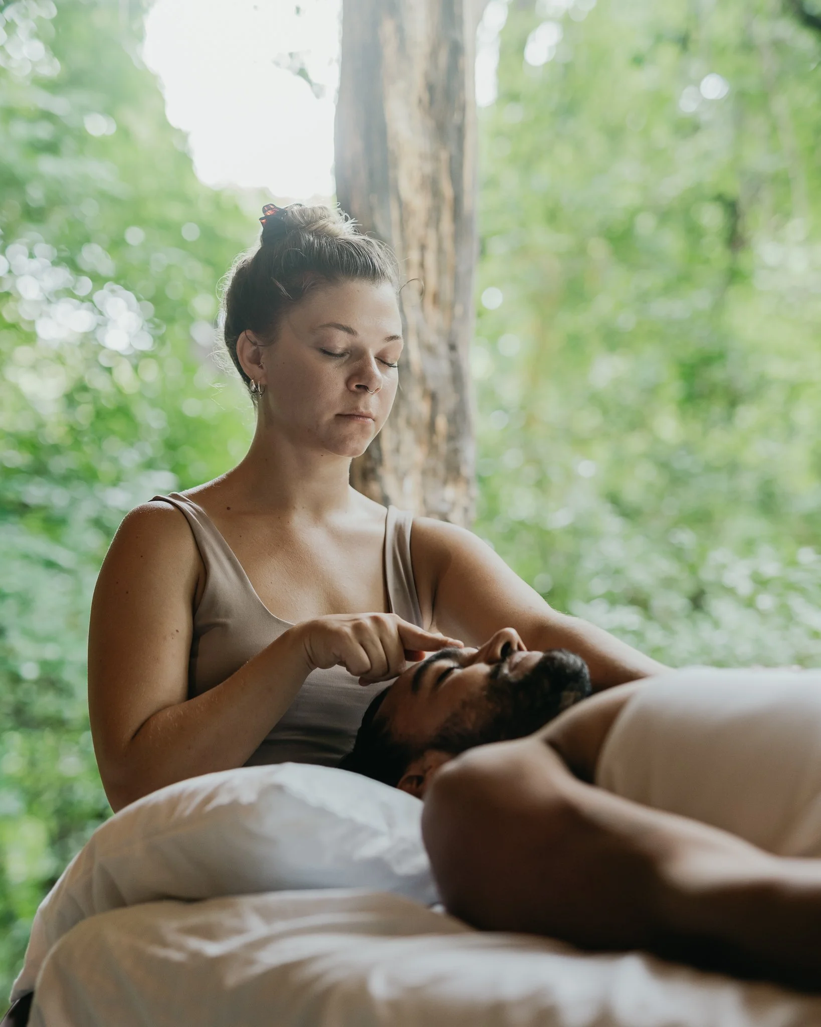 A woman gently touches a man's forehead while he lies on a bed outdoors surrounded by green trees.