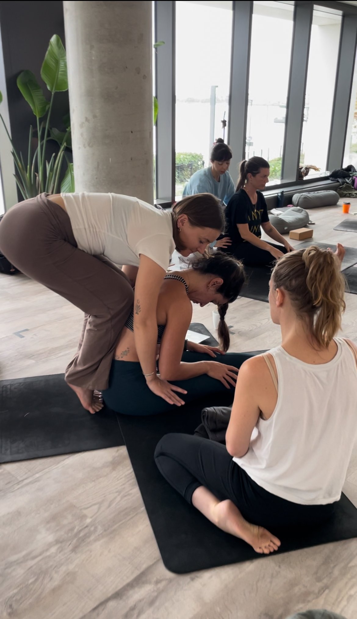 A woman guiding another woman through a yoga pose on a mat during a yoga class in a spacious room with large windows and plants.