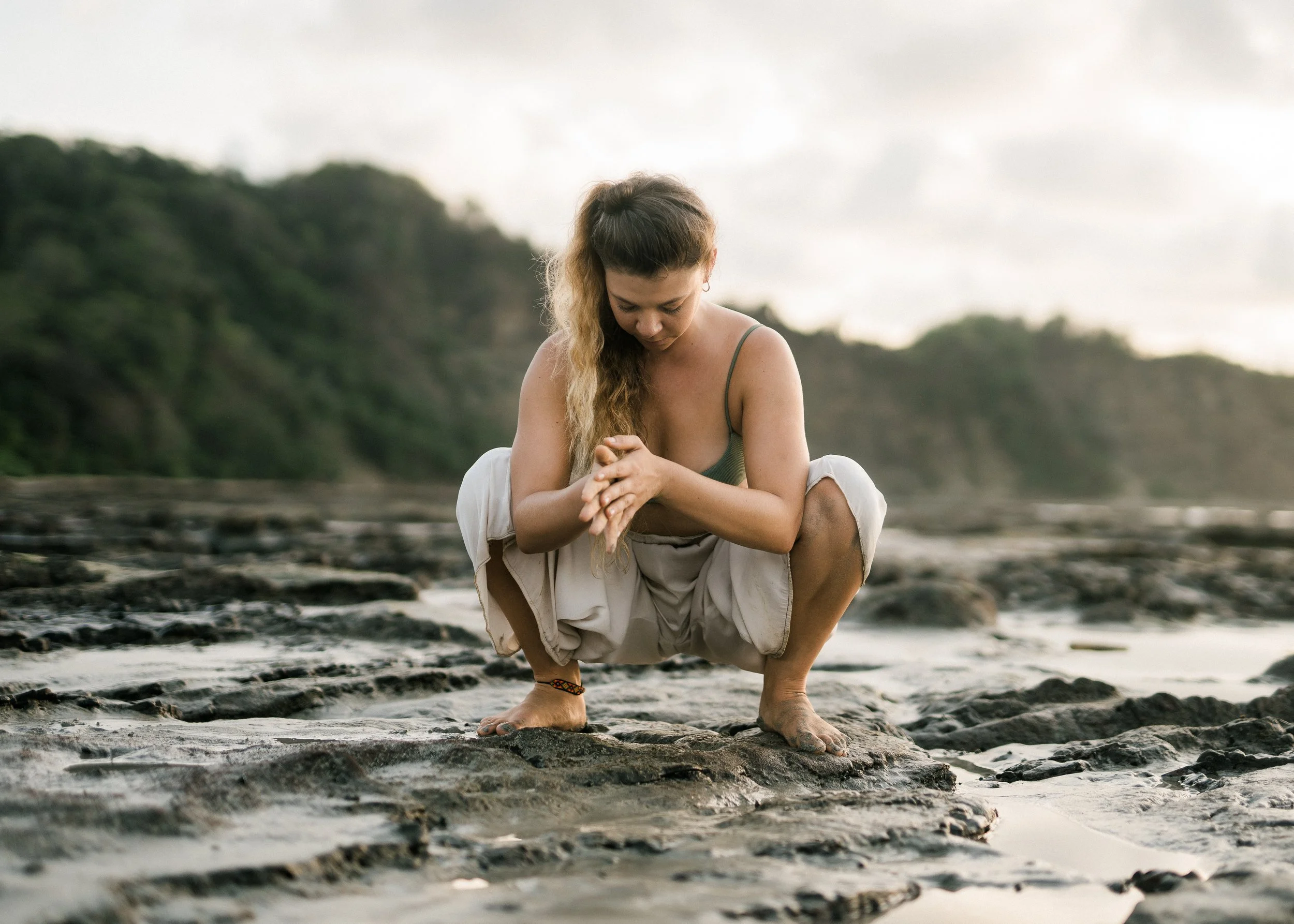 A woman with long hair squatting on a rocky shoreline with water and greenery in the background, looking down with hands clasped.