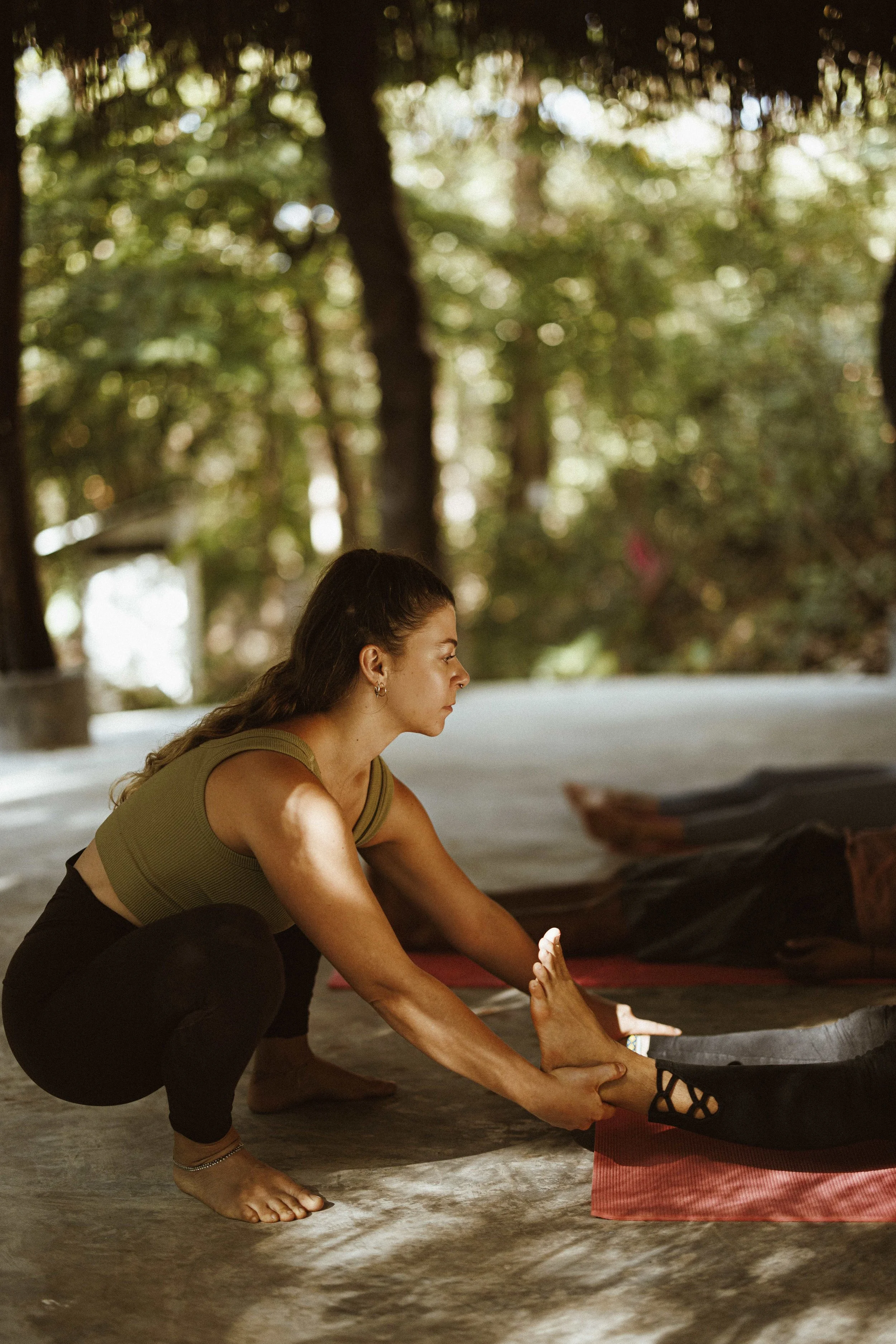 A woman doing yoga outdoors, assisting another person with a stretch or pose on a red yoga mat. Trees and natural surroundings are visible in the background.